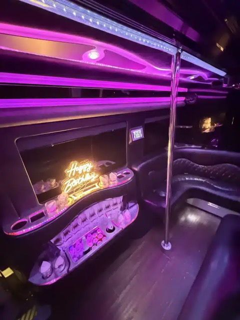 Purple-lit luxury party bus interior with neon sign, leather seating, and chrome pole