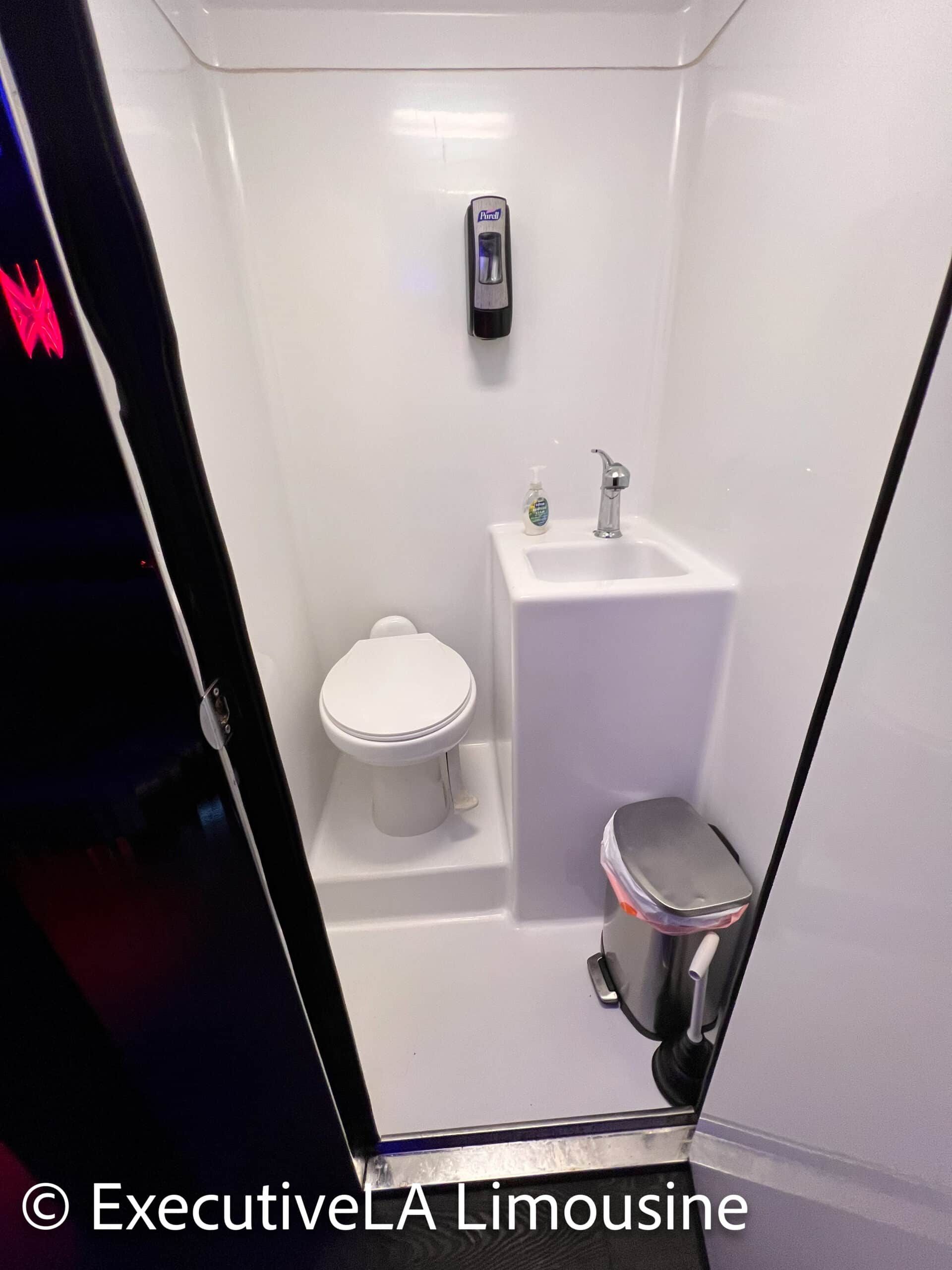 Compact white bathroom with toilet, sink, wall dispenser, and small trash can inside a vehicle or trailer.