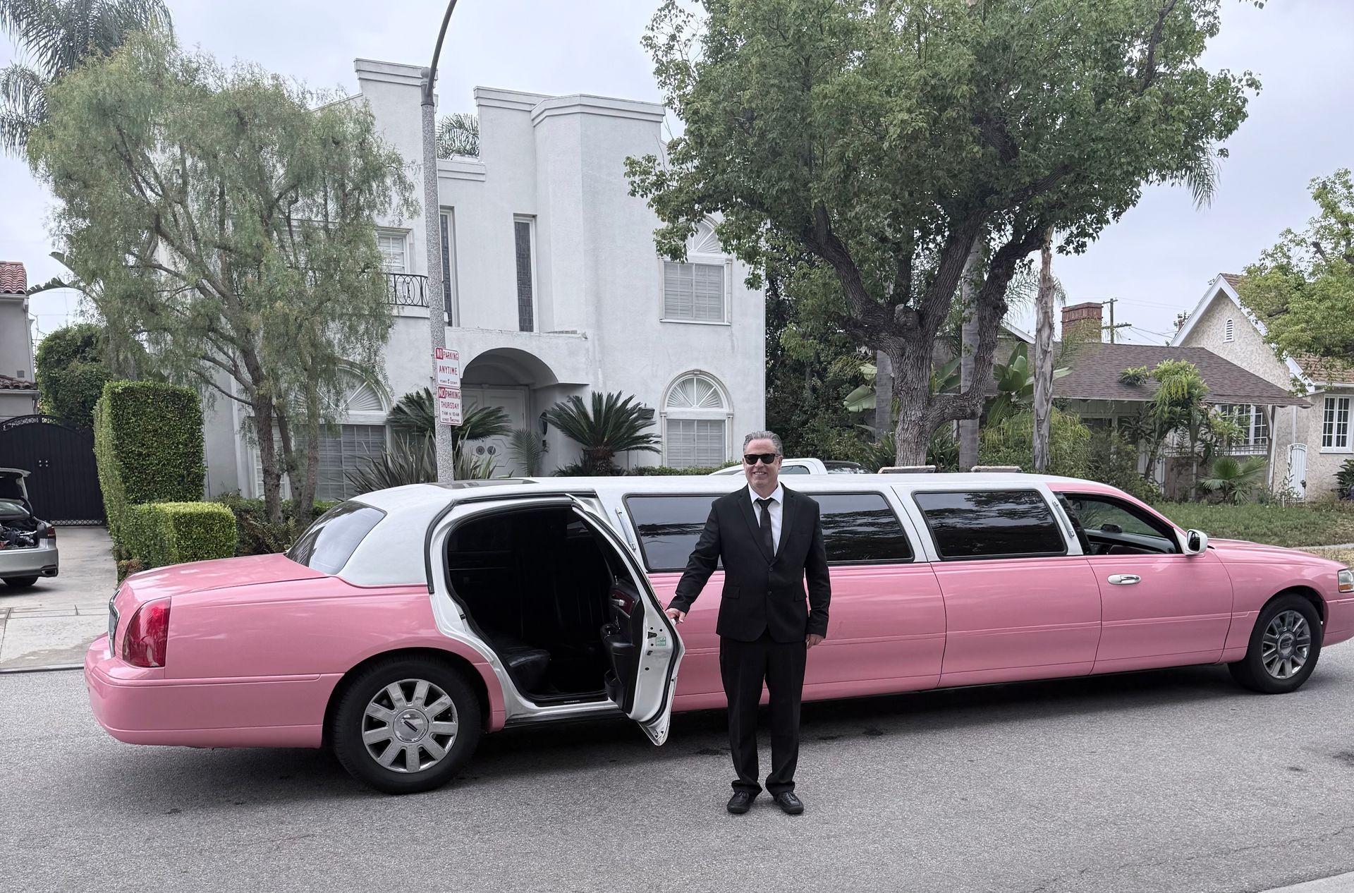 Man in suit standing by a pink stretched limousine with rear door open on a residential street.