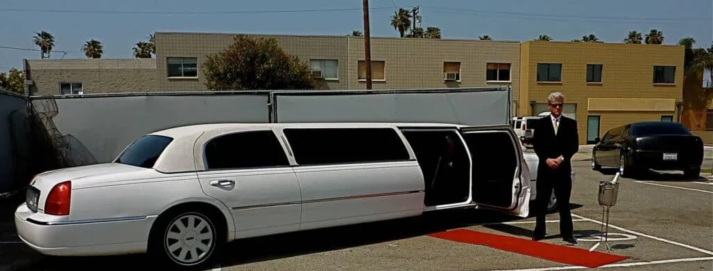 White stretch limousine with open door parked outside a building, with a suited driver standing beside it.