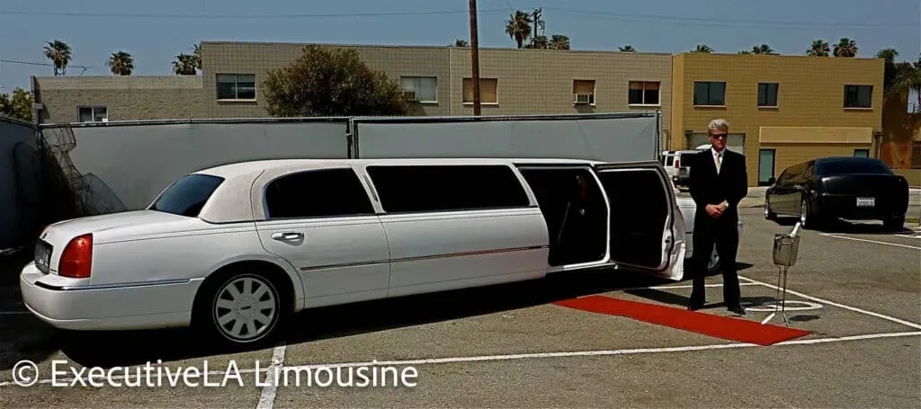 White stretch limousine with open door and red carpet, chauffeur standing outside in a parking lot.