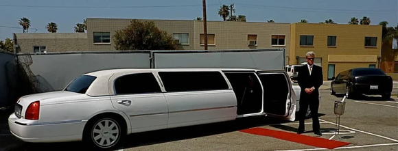 White stretch limousine with open door parked outside a building, with a suited driver standing beside it.