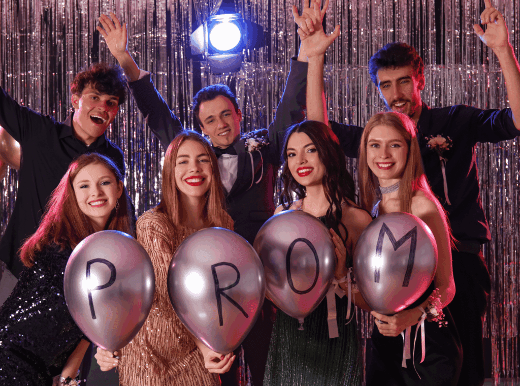 Group of people holding balloons spelling “PROM” at a shiny party backdrop.