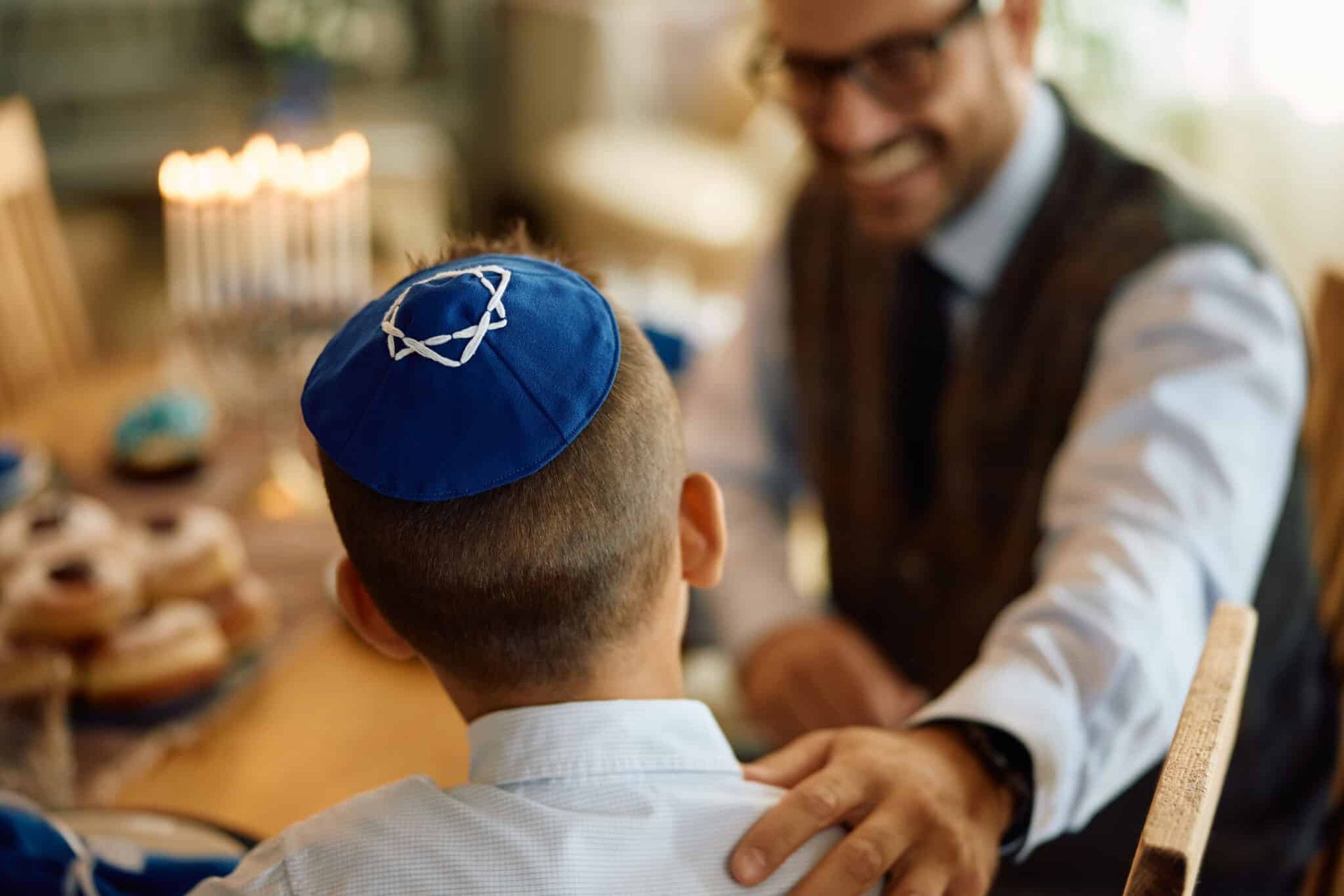 Man wearing a blue kippah sits at a table while a smiling man in glasses touches his shoulder.