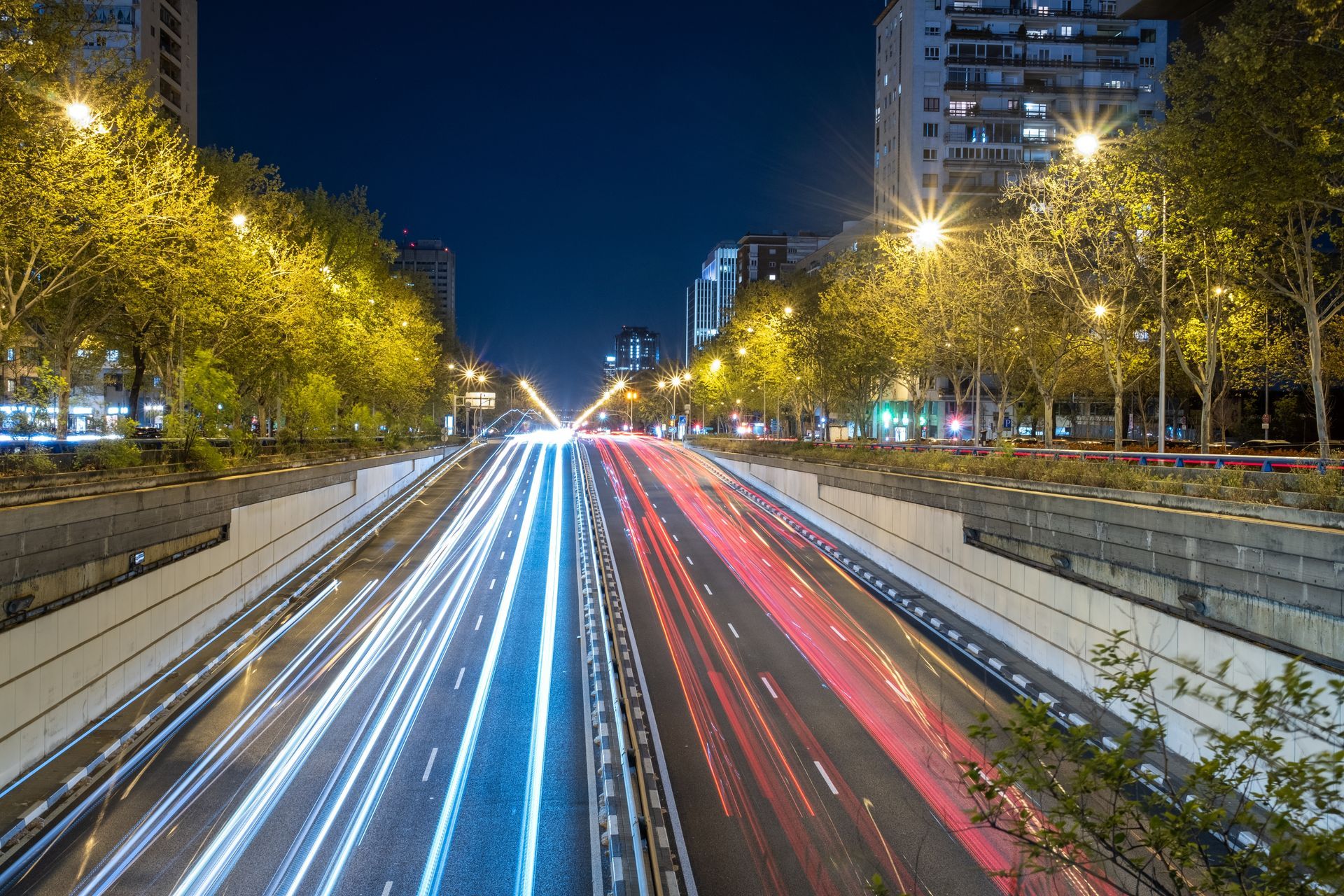Nighttime city boulevard with long-exposure white and red traffic light trails between tall buildings