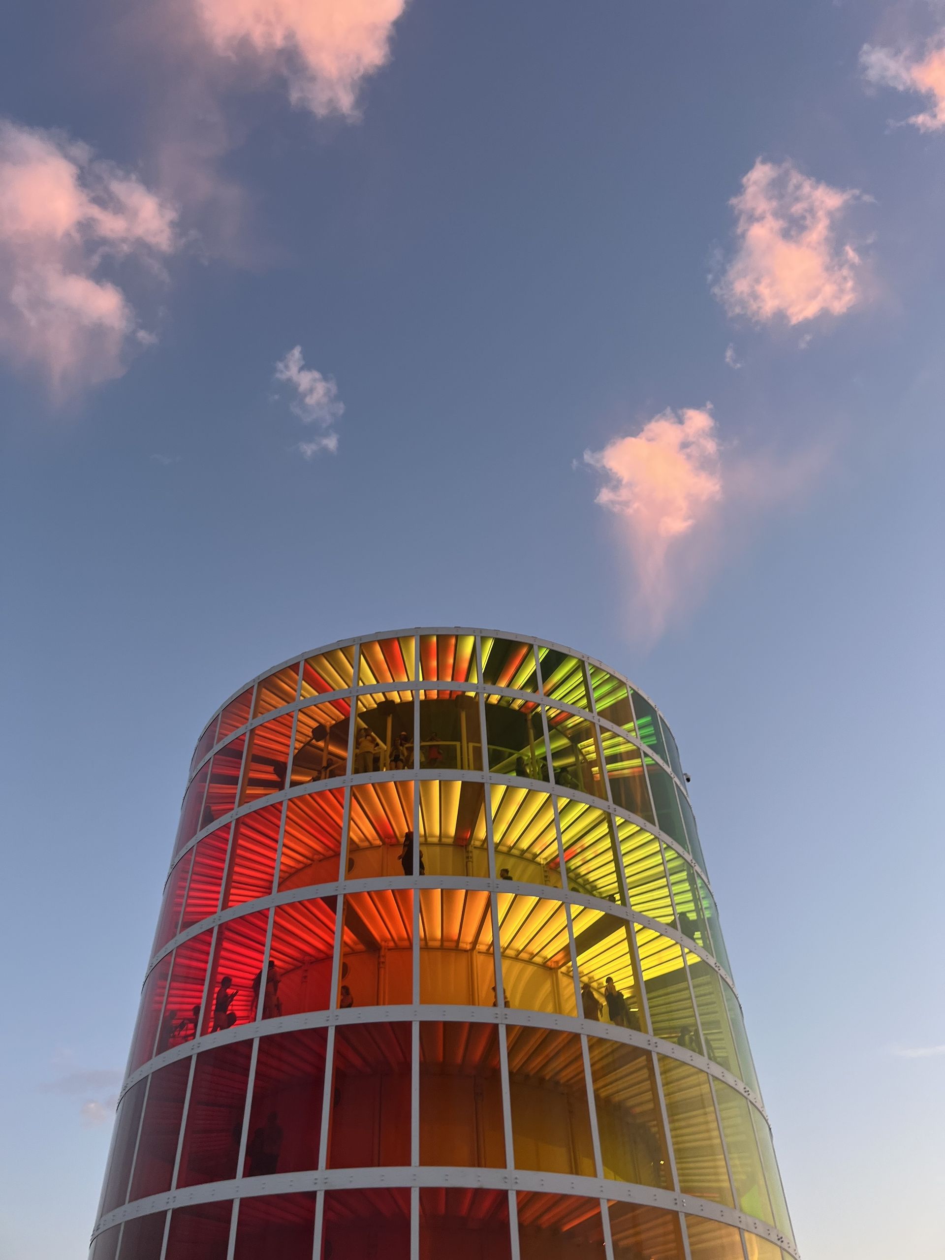 Colorful cylindrical tower lit red, orange, and yellow against a blue sky with pink clouds