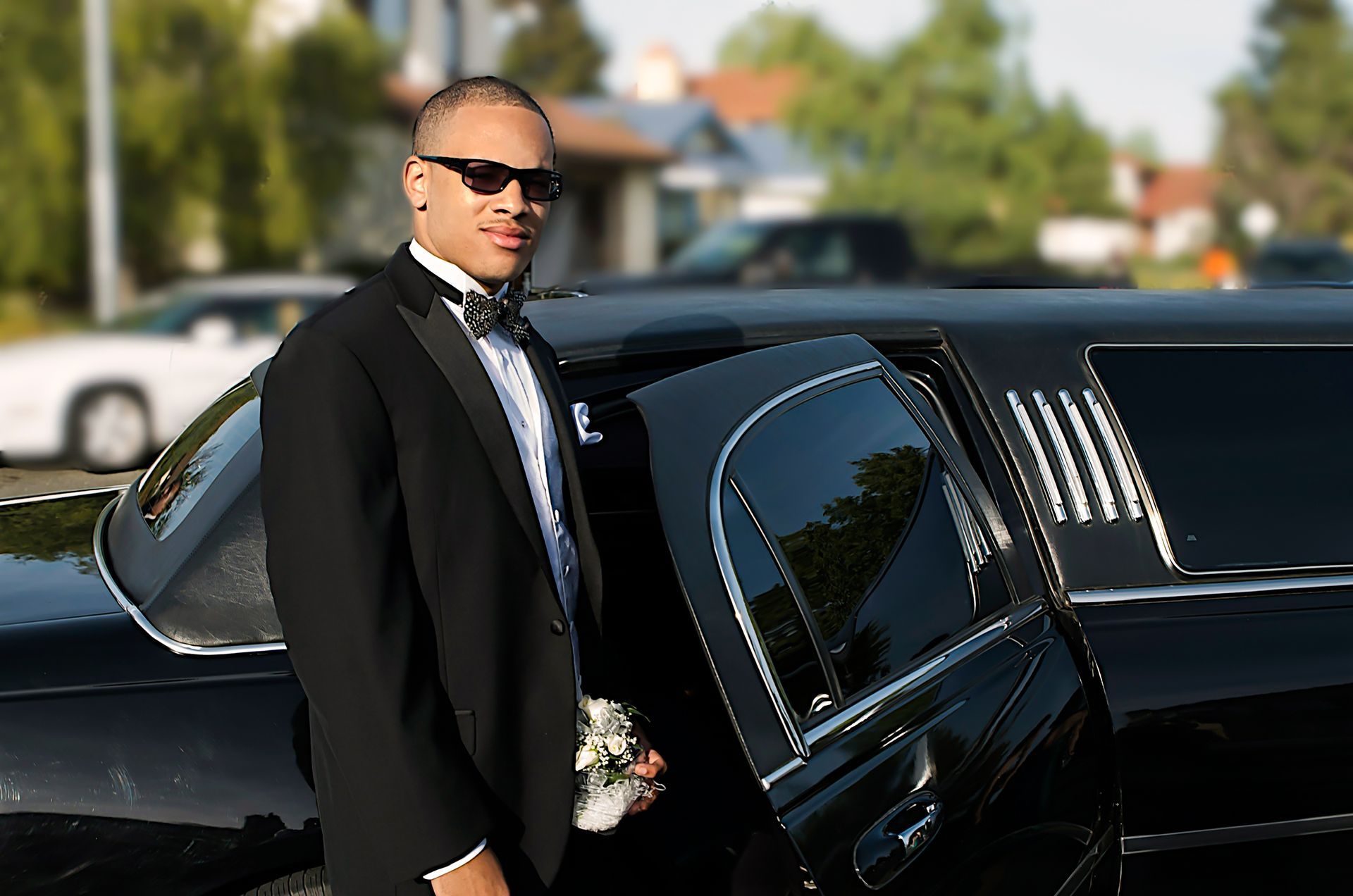 Man in a black suit standing beside a black limousine outdoors on a sunny day