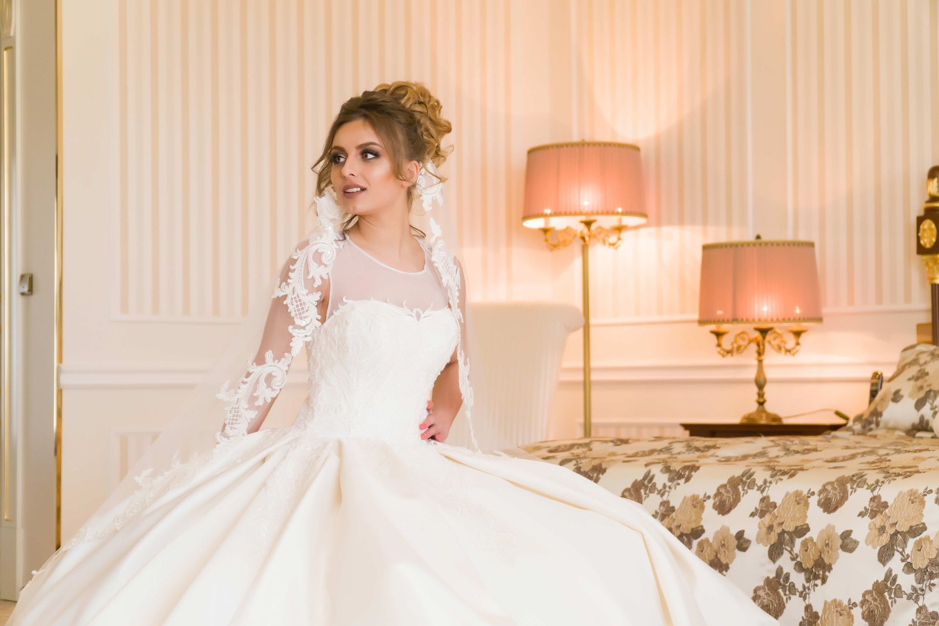 Bride in a white lace wedding gown standing in an elegant bedroom beside a floral bed and pink lamp.