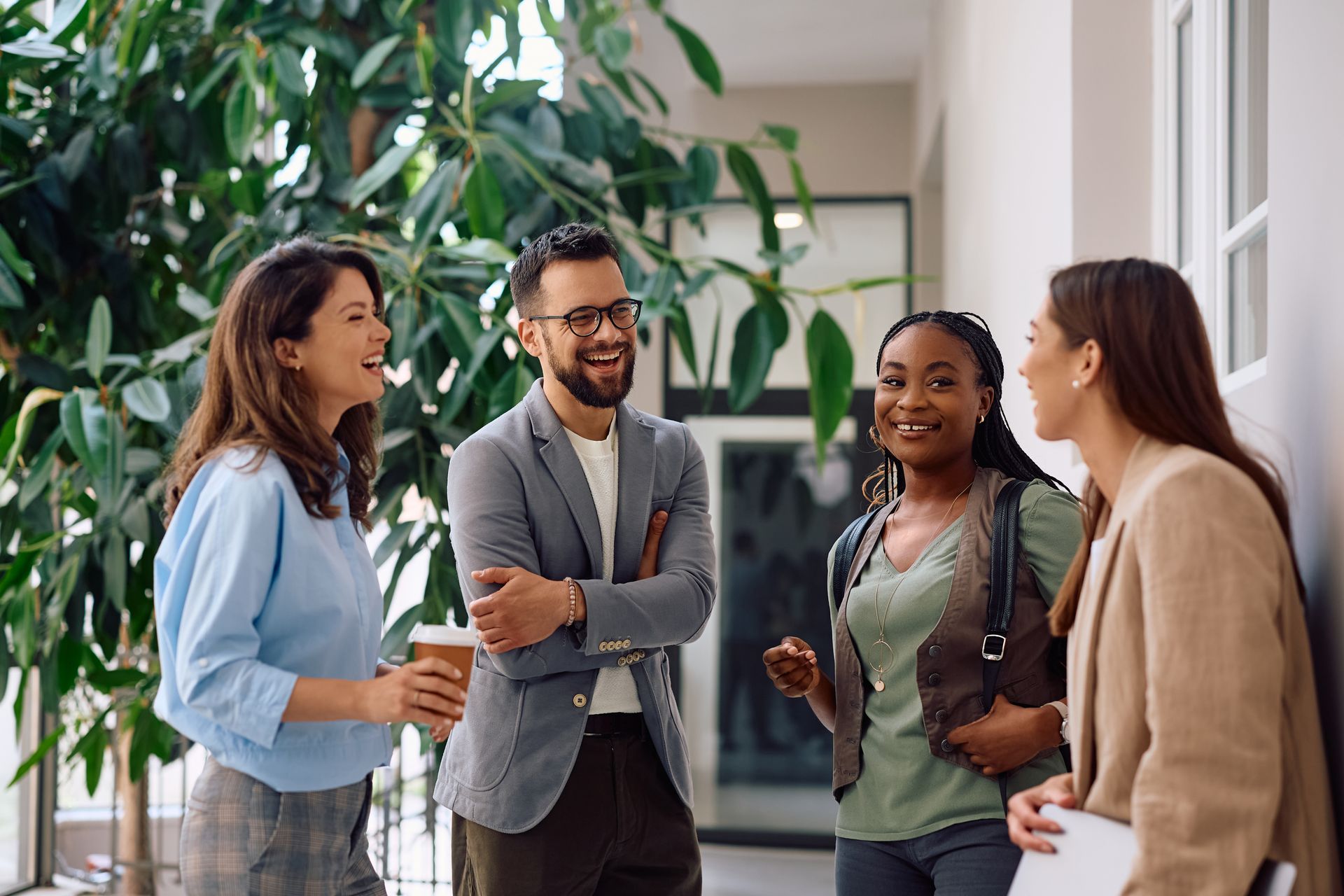 Four coworkers chatting and smiling in a bright office hallway, holding coffee and papers.