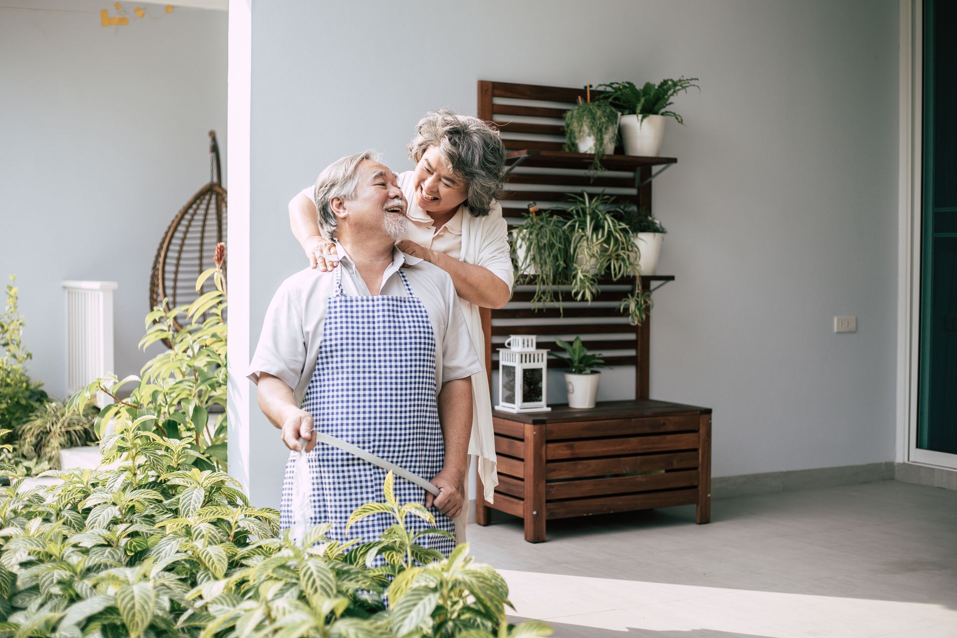 Elderly couple smiling in a bright room beside a plant shelf and hanging chair.