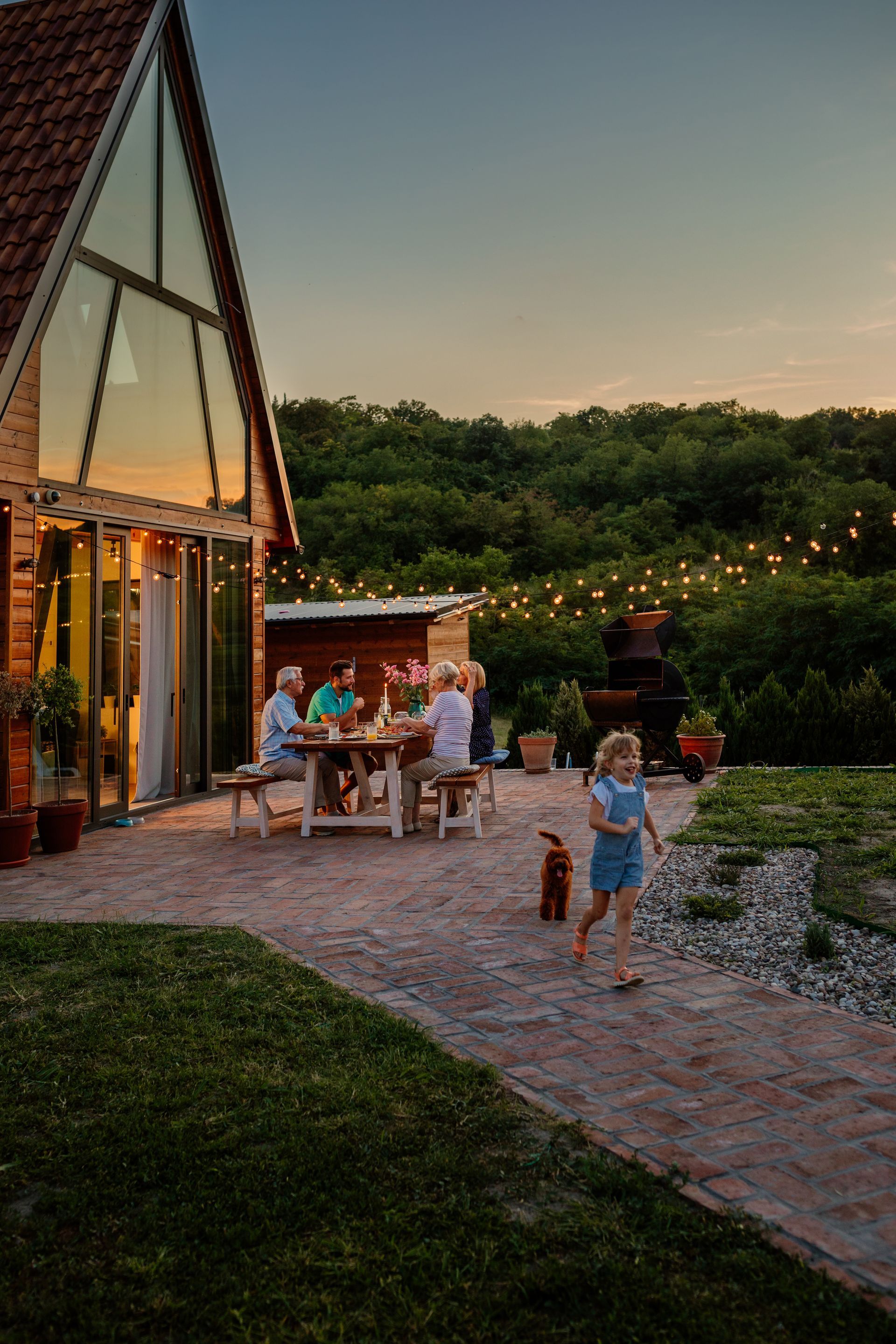 People dining outdoors by a lit glass house at sunset, with children on a stone path and string lights beyond.