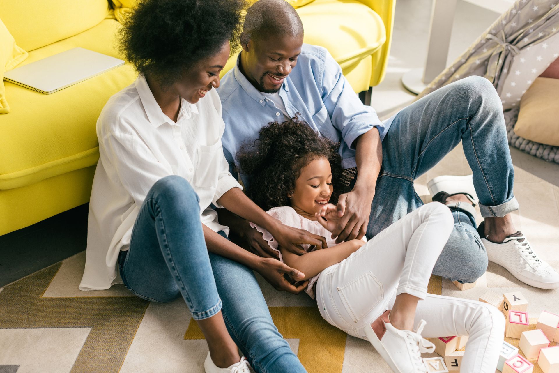 Family sitting on the floor by a yellow couch, smiling and playing with a child among scattered toys.