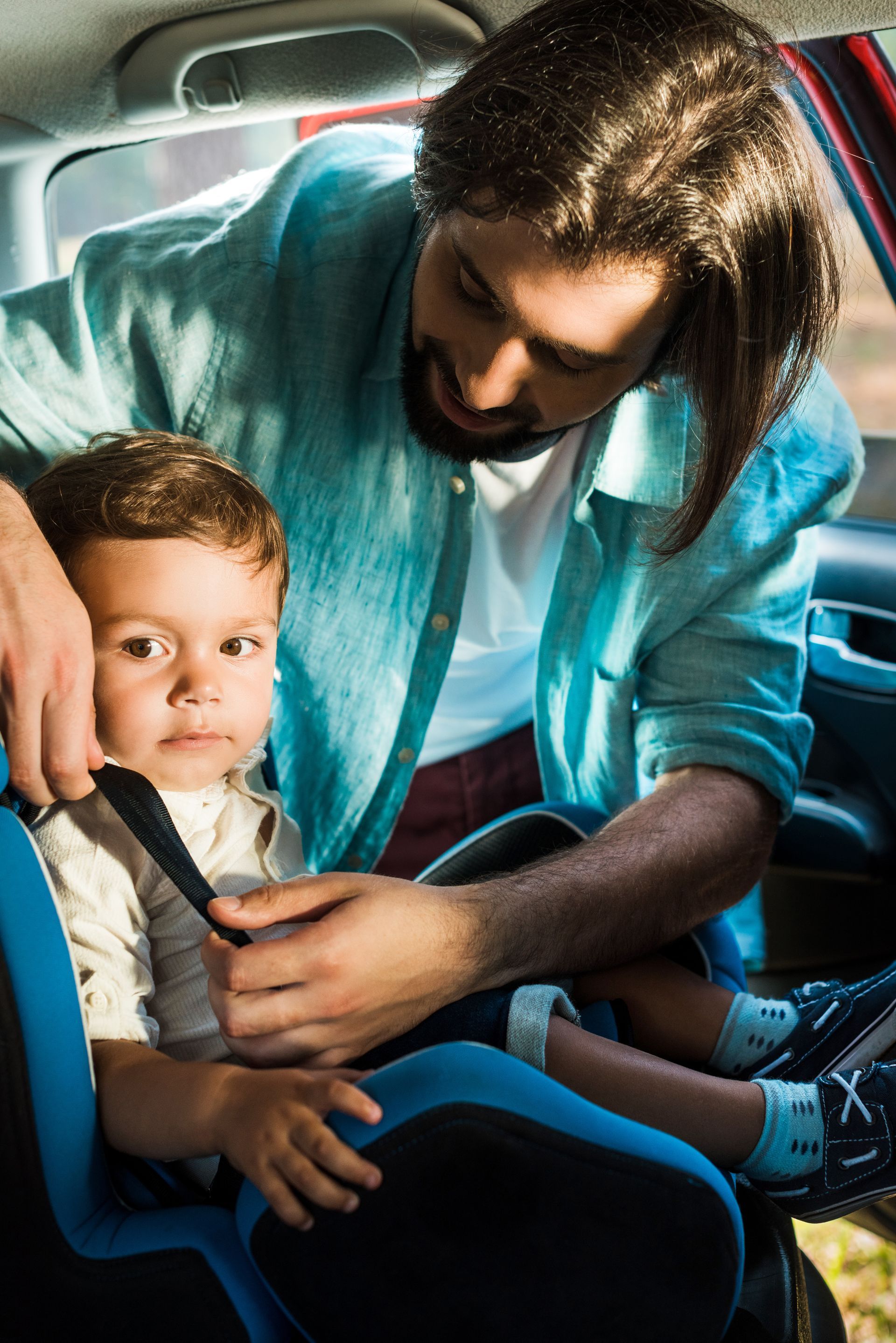 Adult buckling a child into a car seat inside a car, both wearing seat belts.