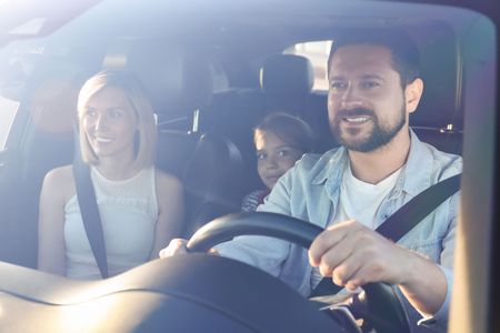 Three people smiling in a car, with the driver at the wheel and bright sunlight through the windshield