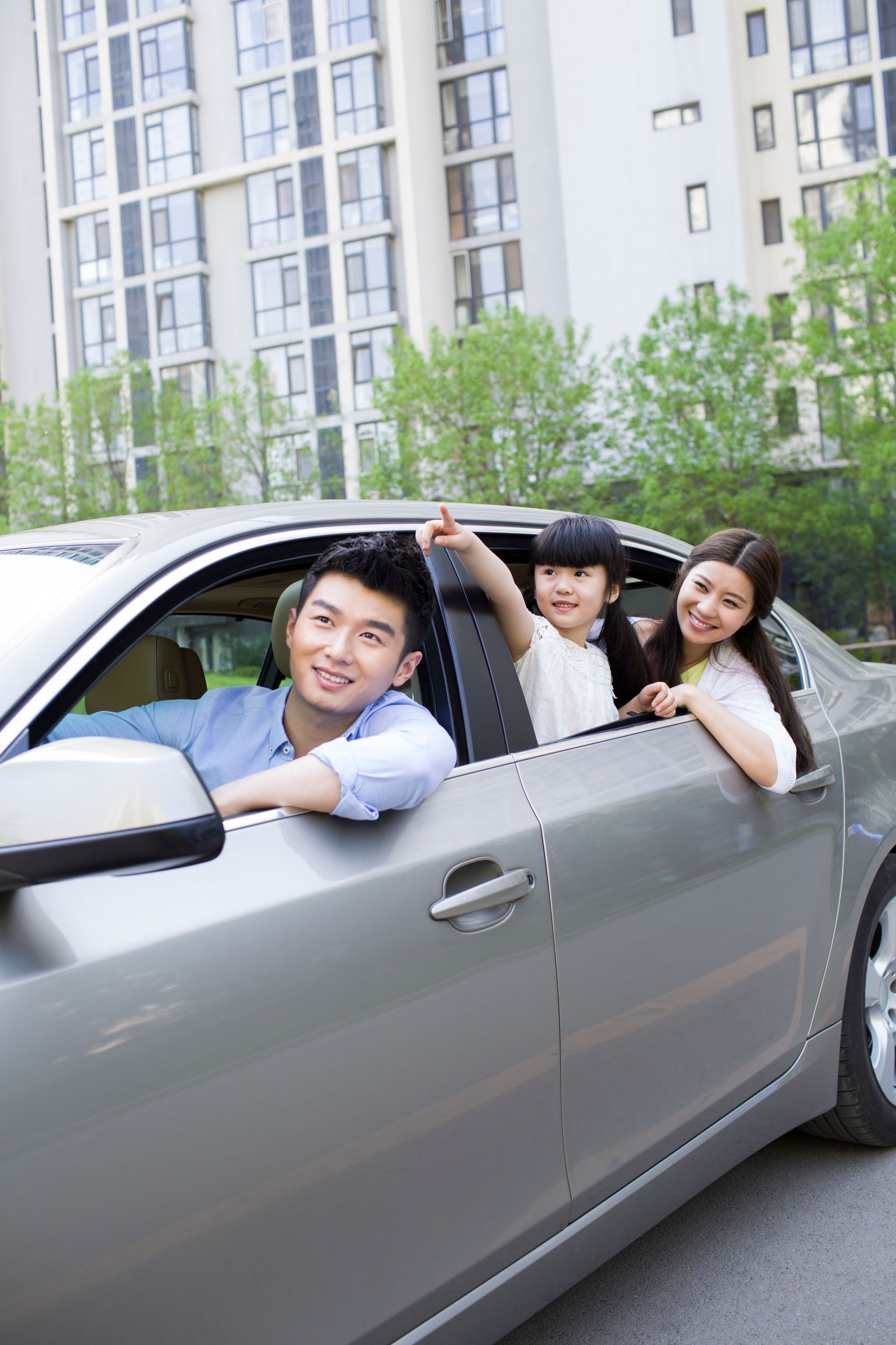 Three smiling people lean out of a silver car parked beside apartment buildings.