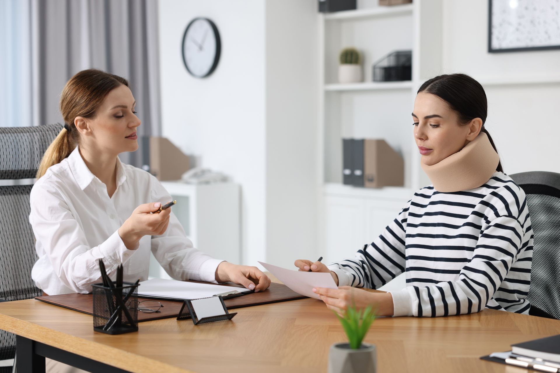 Two women discussing documents at a desk in a bright office, one in a white shirt and one in a striped top.