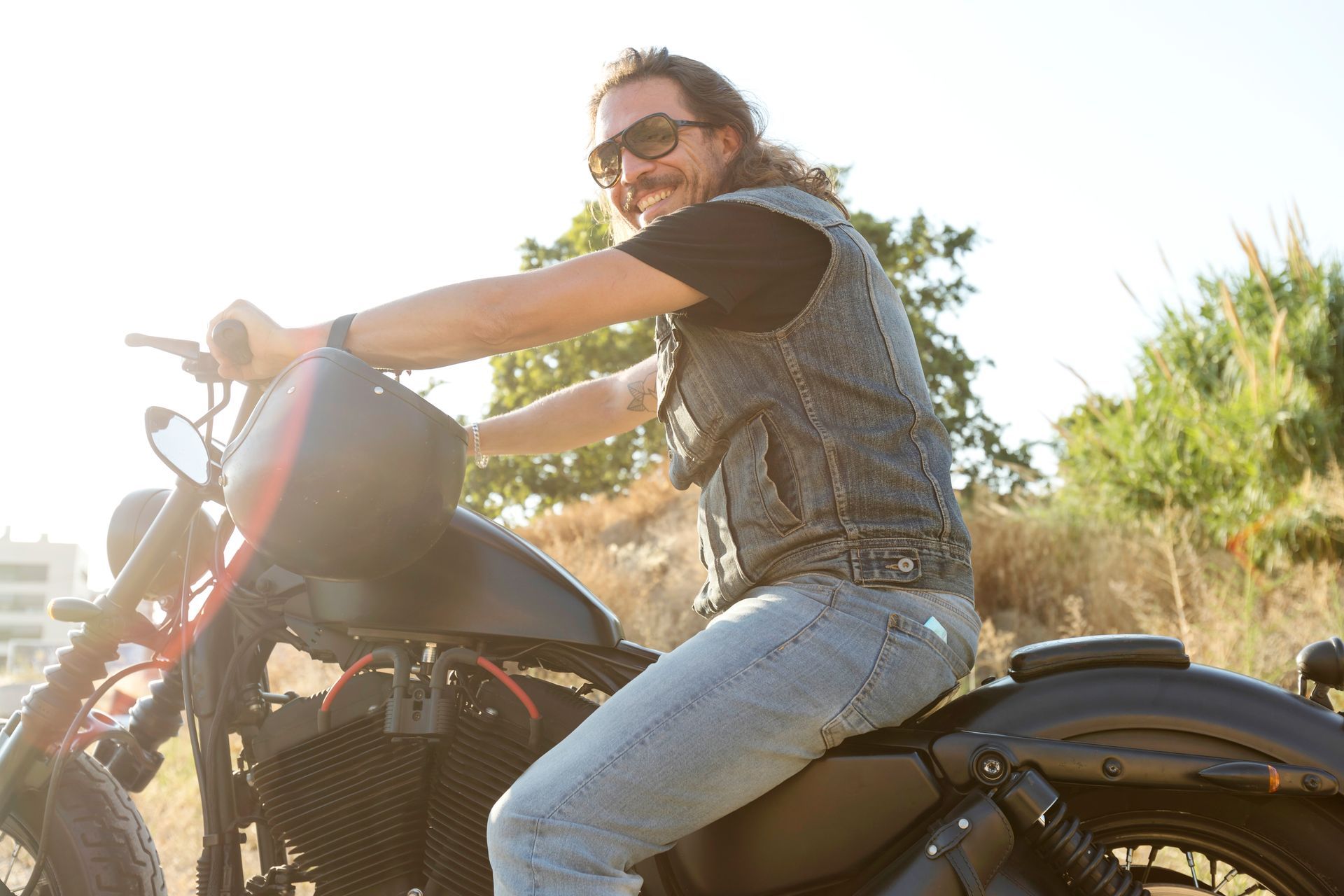 Person on a motorcycle in a sunlit outdoor setting, wearing sunglasses and a vest.