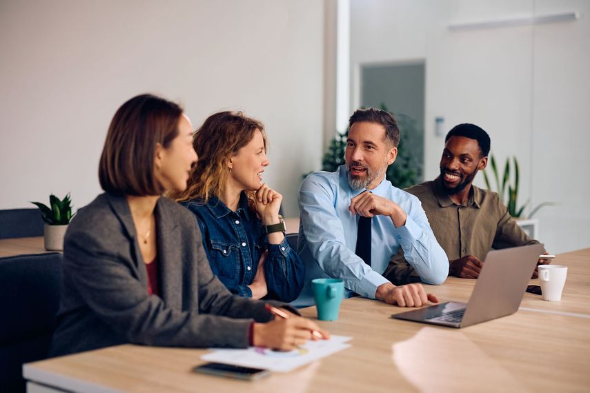 Colleagues in a meeting around a table, talking and using a laptop in a bright office.