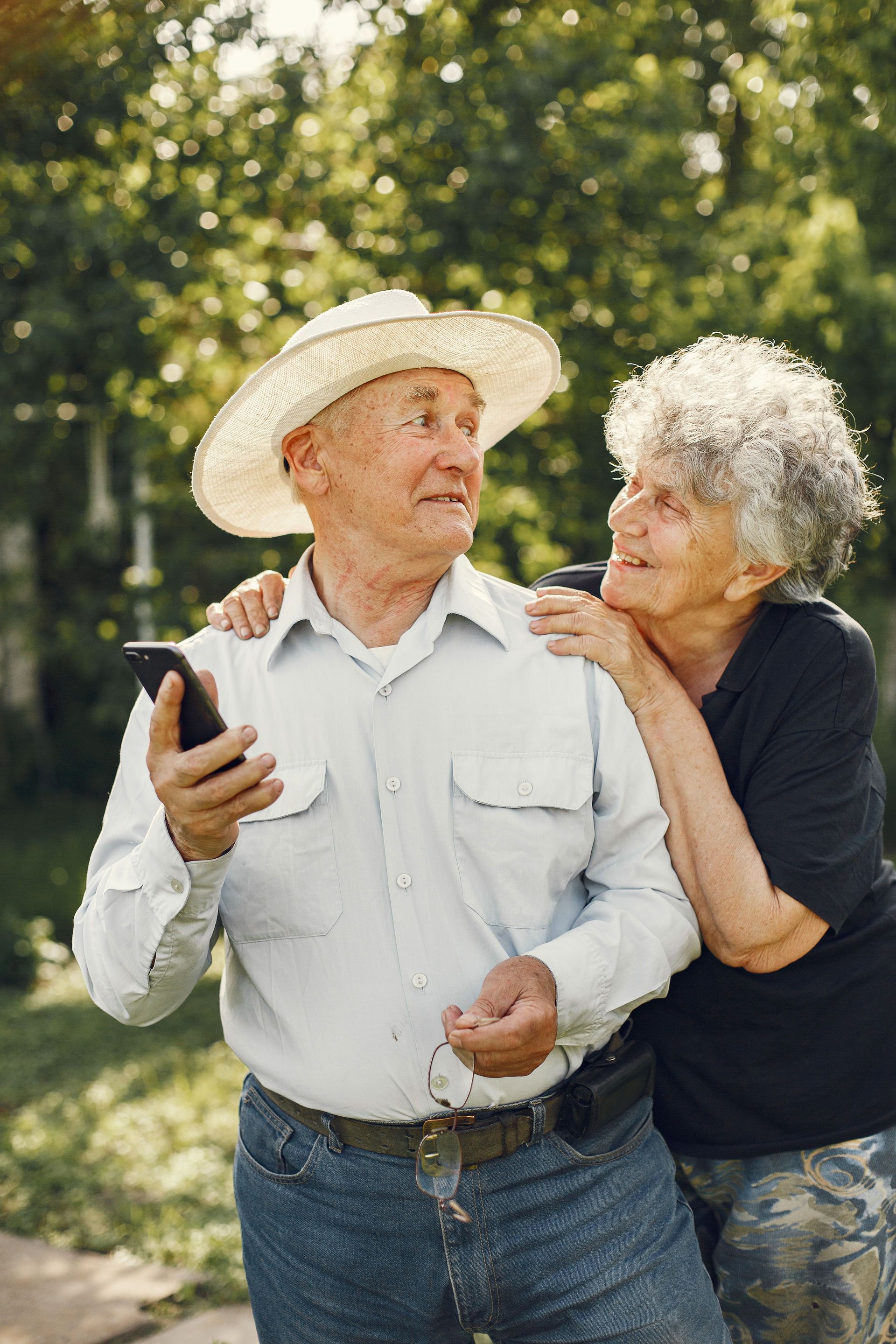 Two people standing outdoors in a sunny garden, one holding a phone while the other leans in to look at it.