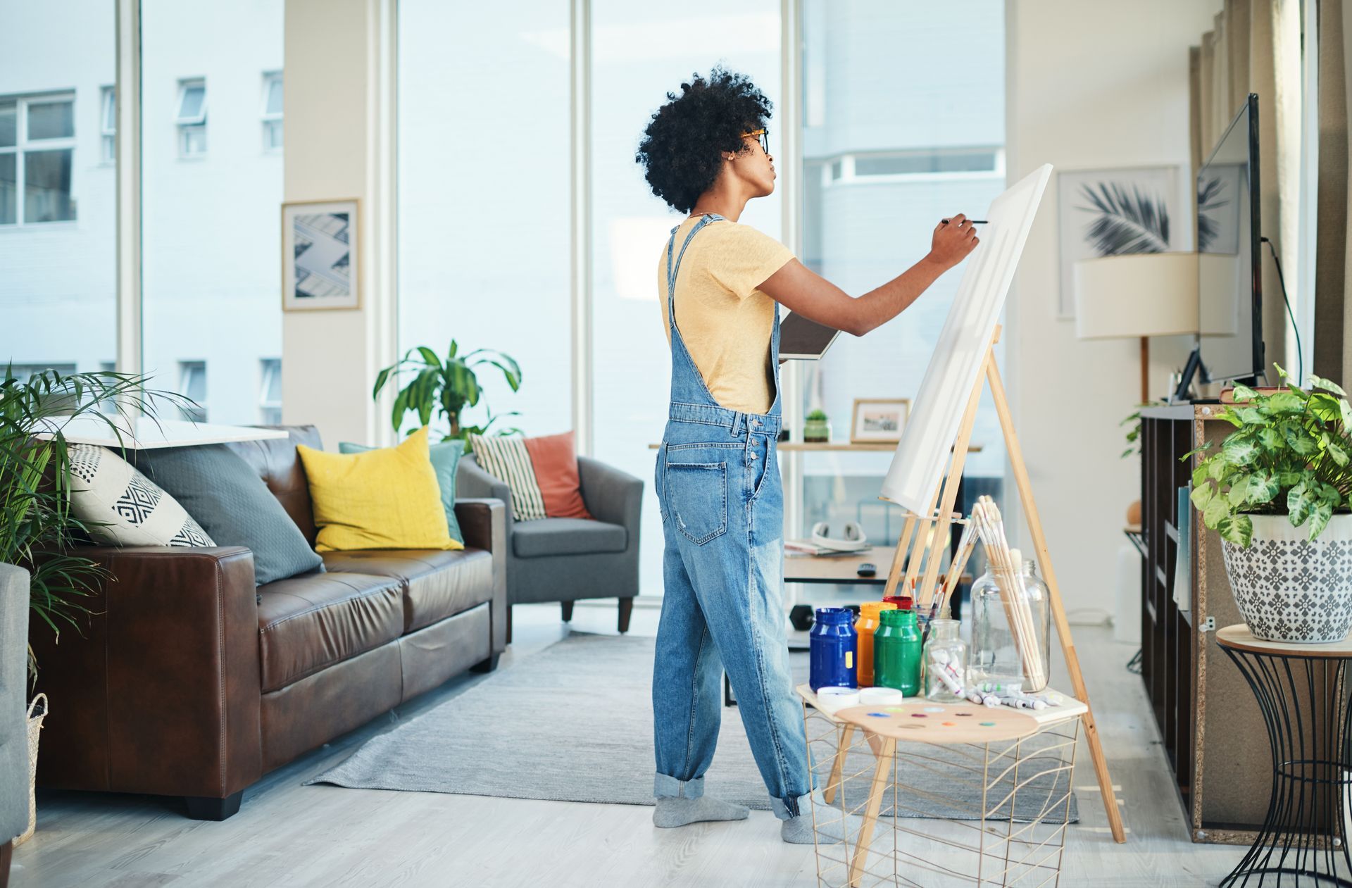 Person painting a wall in a bright living room with a couch, plants, and a ladder