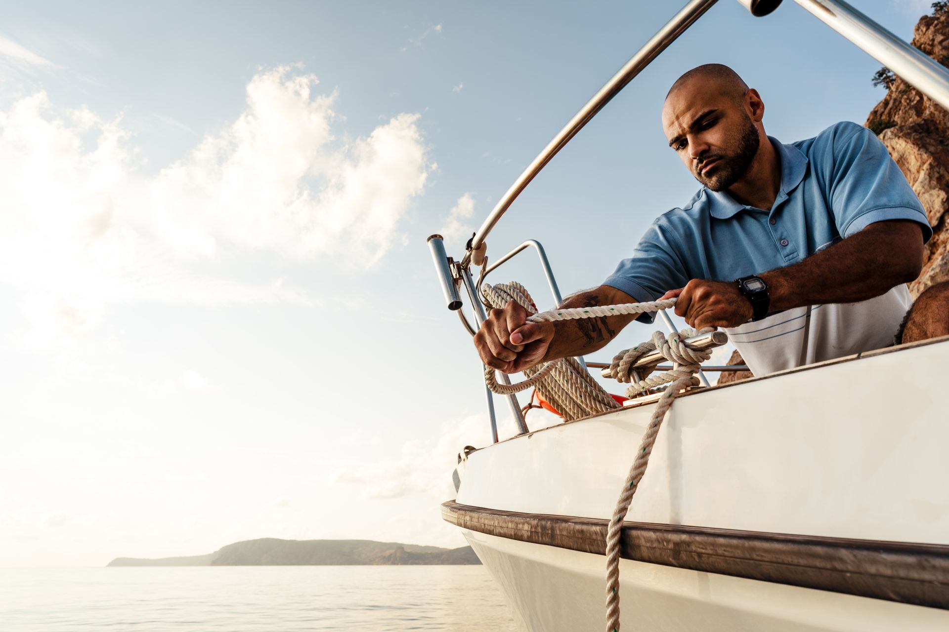 Man in blue shirt on a sailboat, adjusting ropes on the deck near calm water at sunset