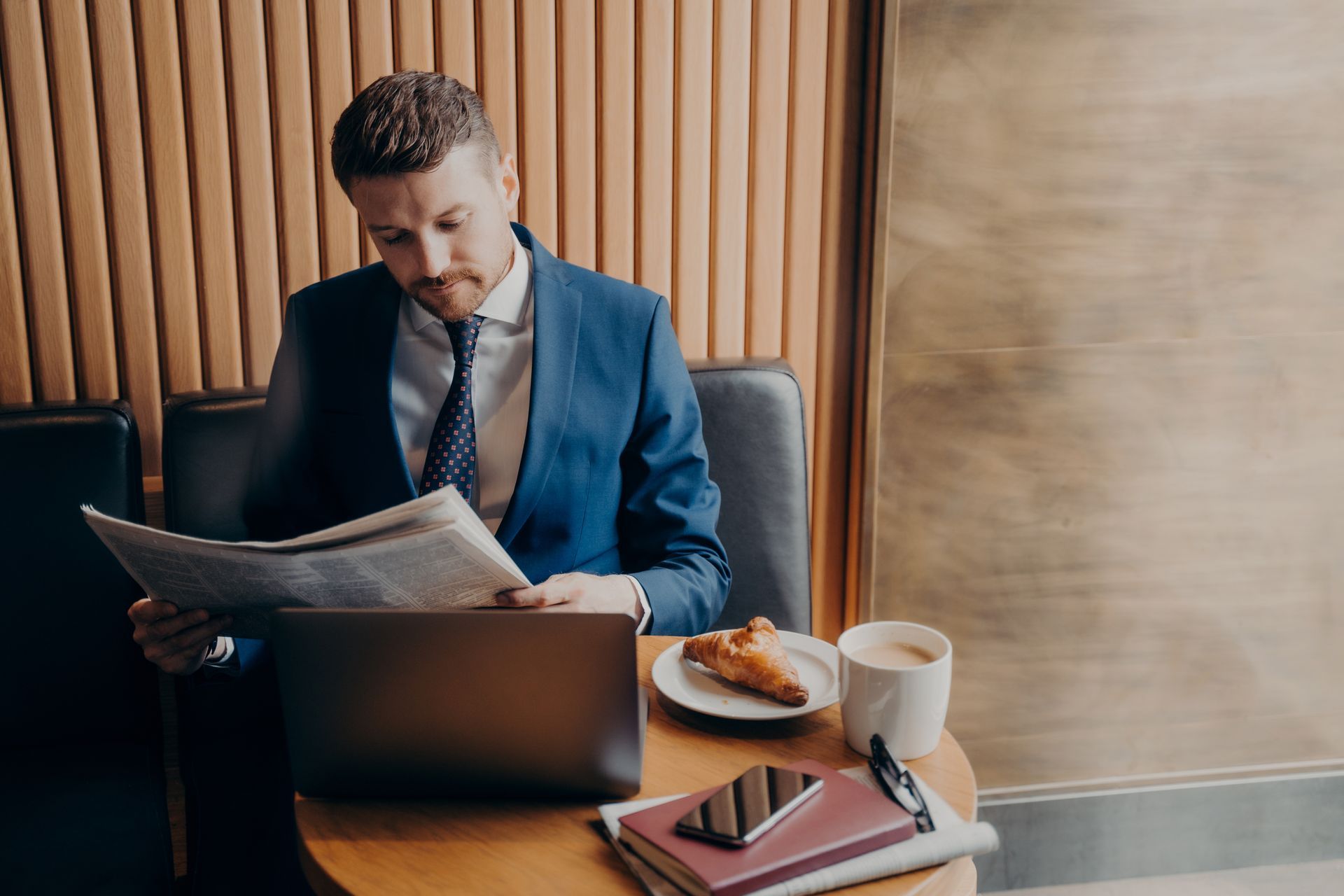 Man in a blue suit reading a newspaper at a café table with a laptop, coffee, and pastry