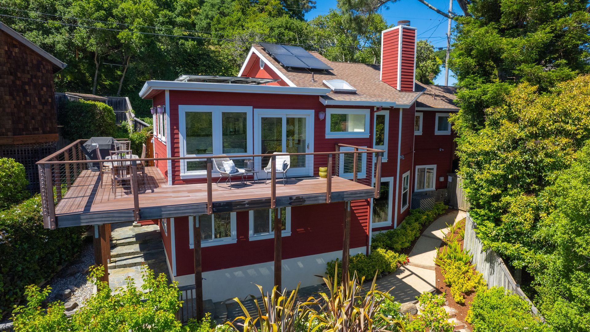 Red house with a rooftop deck, large windows, and a chimney surrounded by trees under a blue sky