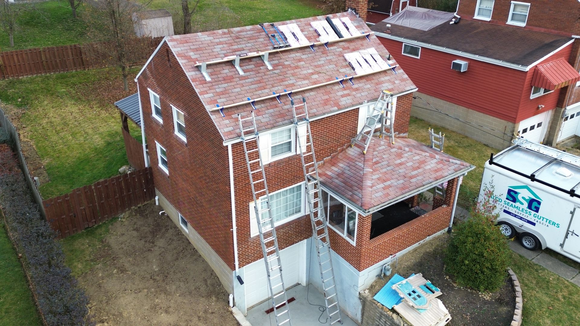 A two-story house with ladders and roofing work in progress; red brick siding and brown roof.