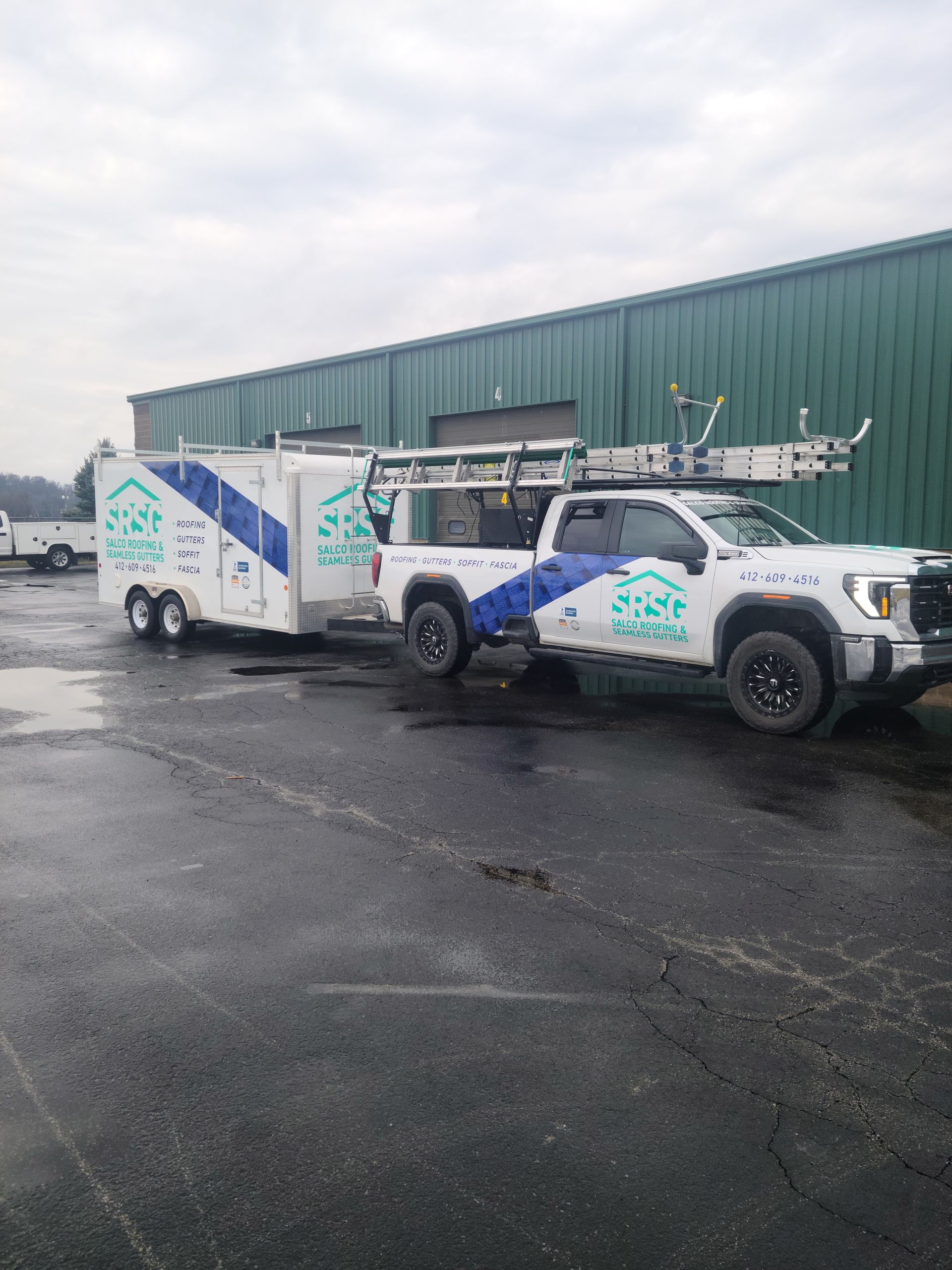 White truck with trailer, both labeled with a company logo, parked in front of a green building.