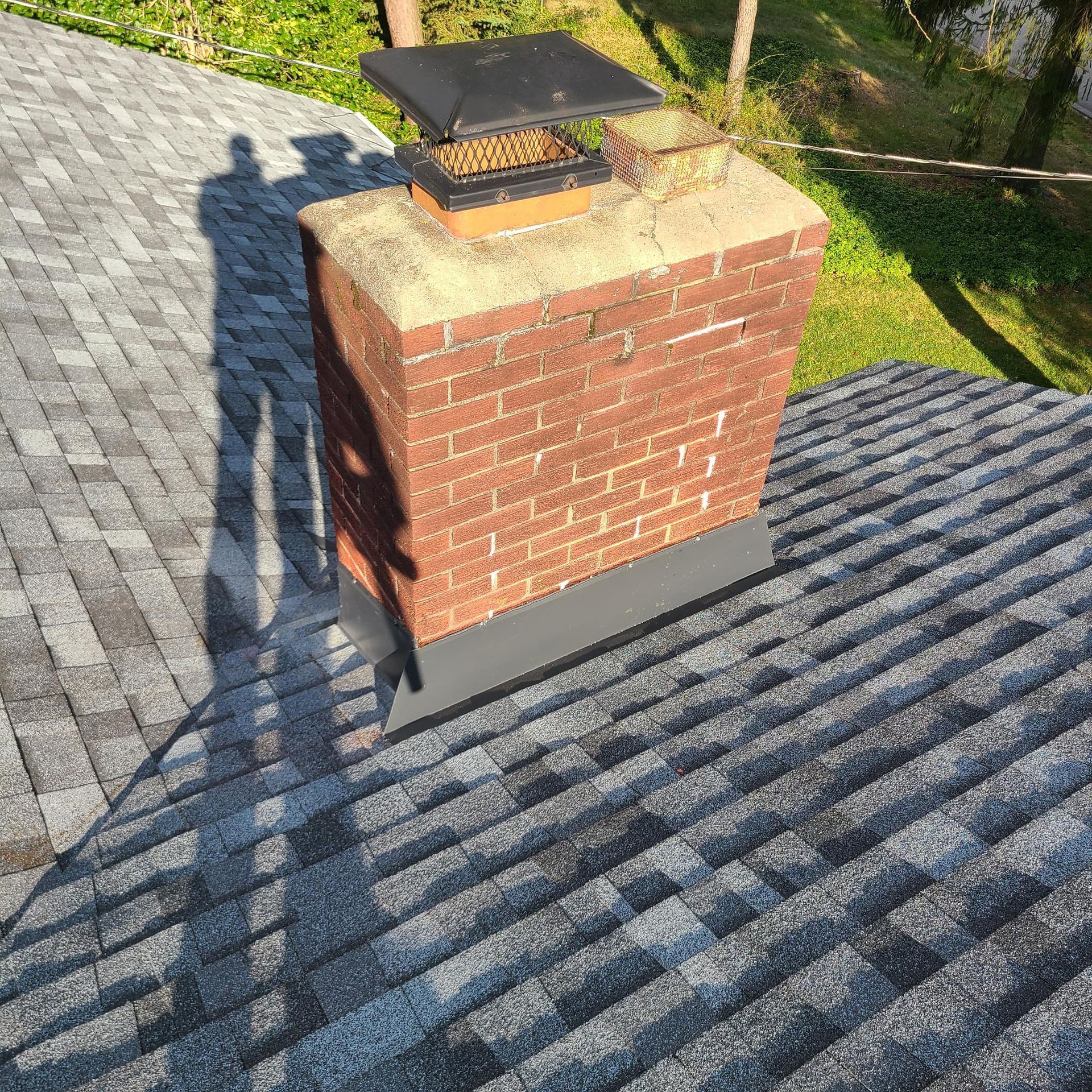 Chimney on a gray shingle roof, with metal flashing and a black cap. Shadow of a person present.