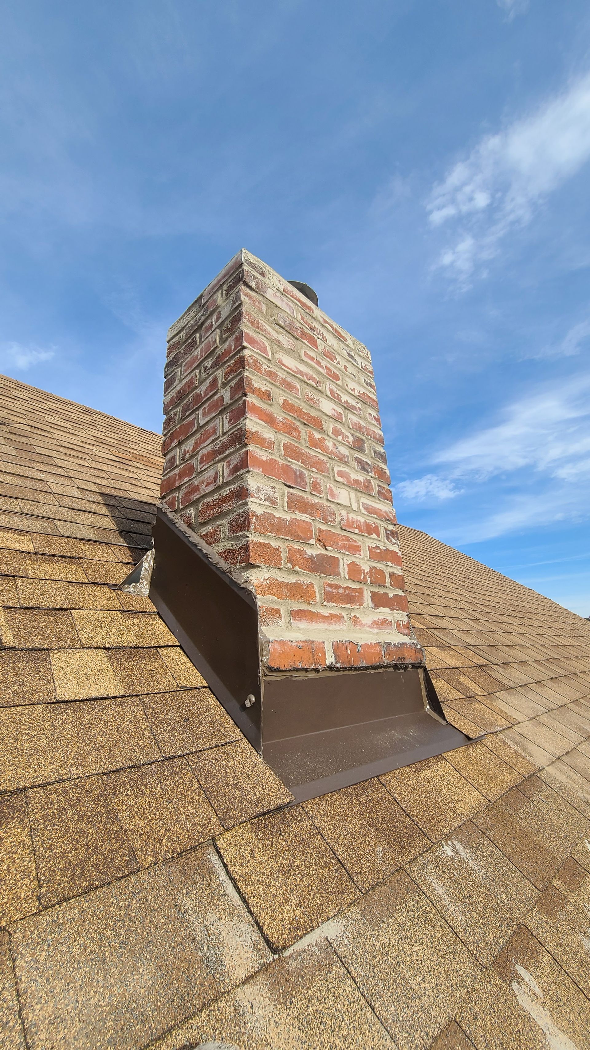 Brick chimney on a shingled roof with metal flashing against a blue sky.