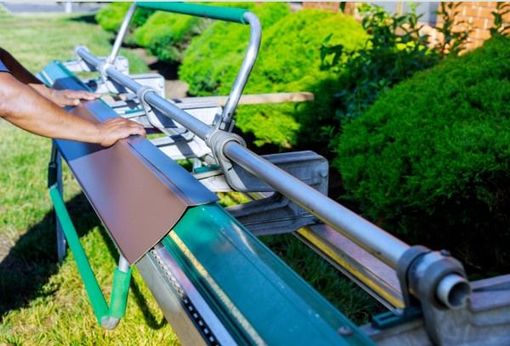 Person using a metal bending tool to shape a brown metal piece, green tool frame, lush green bushes in the background.