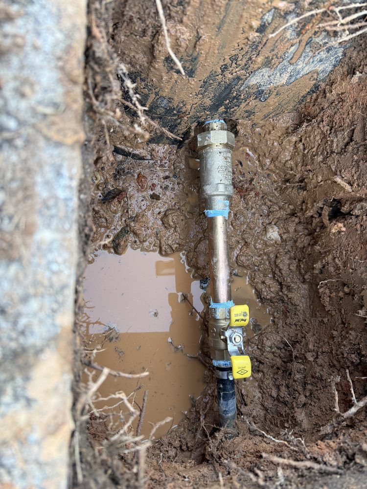 A buried metal pipe with fittings in a muddy trench beside exposed roots and soil.