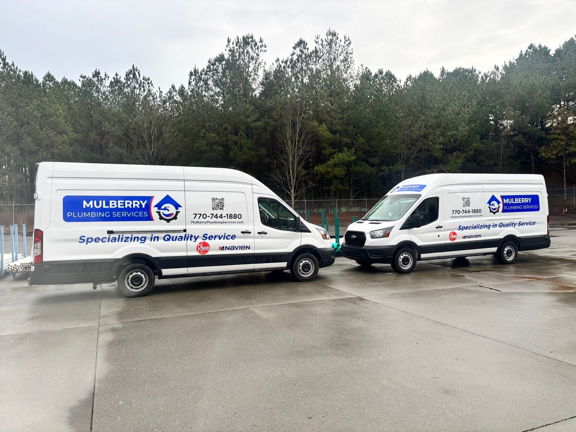 Two white cargo vans with blue logos parked on a wet road by trees
