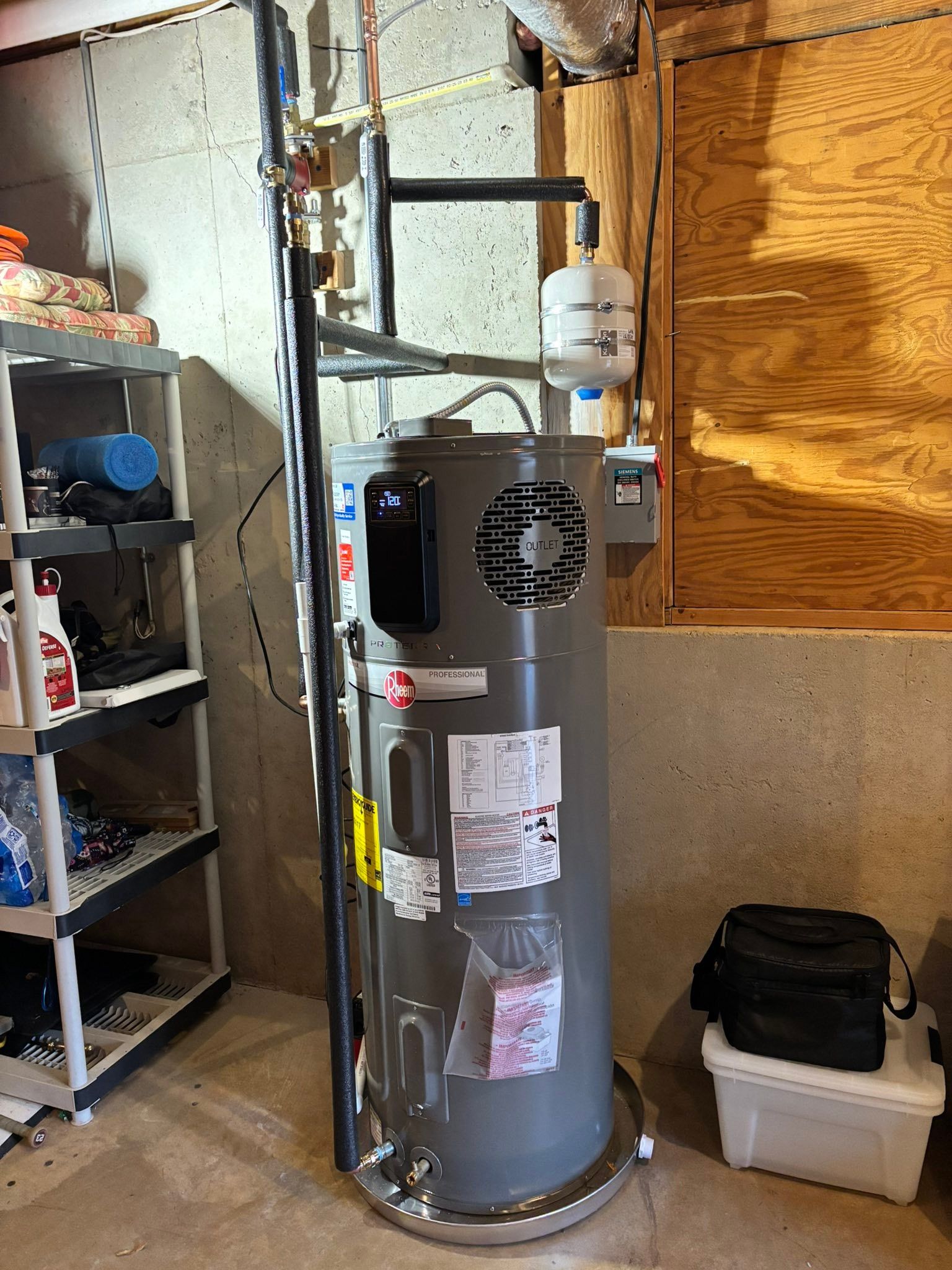 Basement water heater beside shelves and a plywood wall, with storage bins nearby.