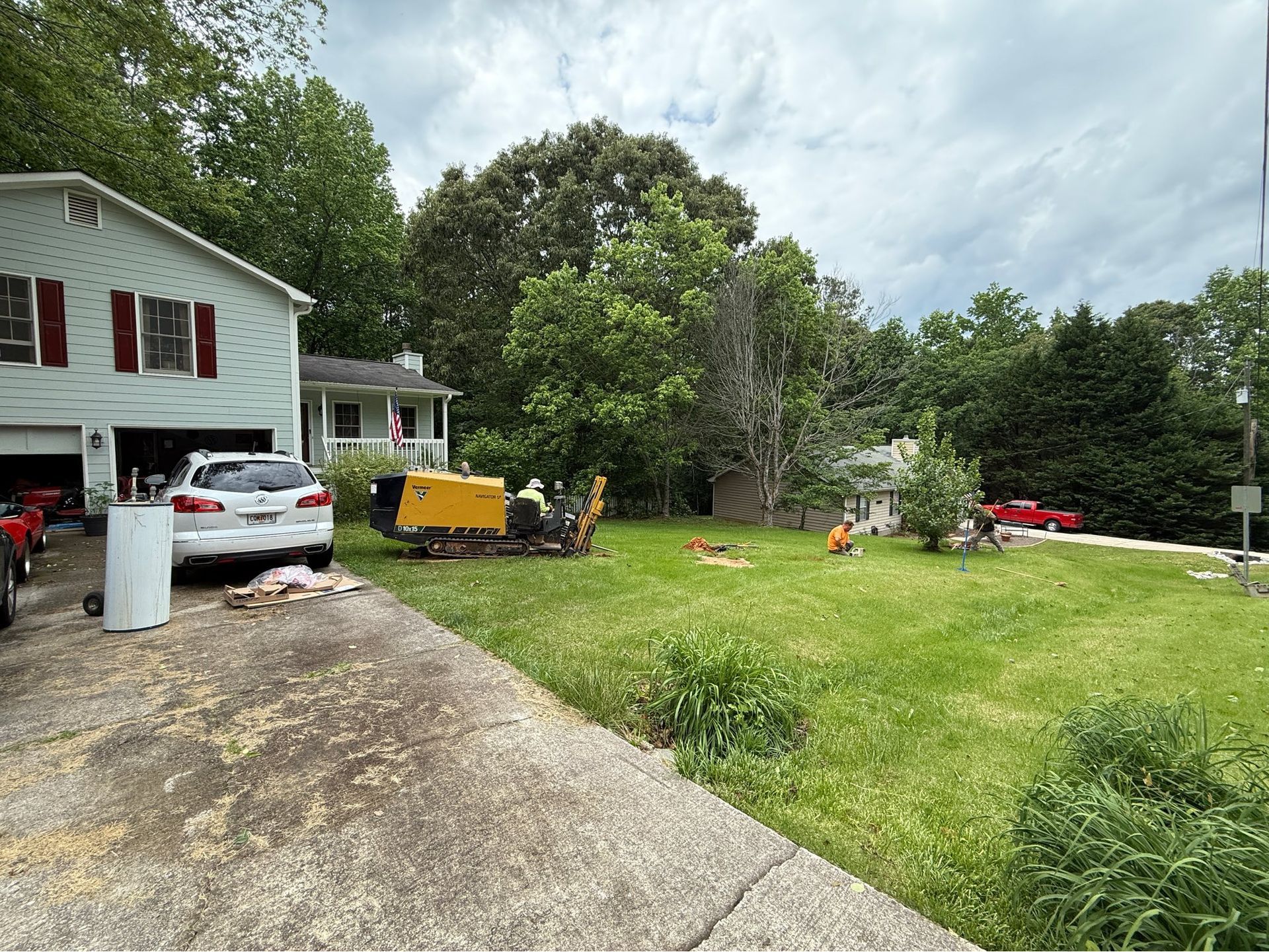 Suburban yard with a white house, parked cars, a yellow machine, and trees under a cloudy sky