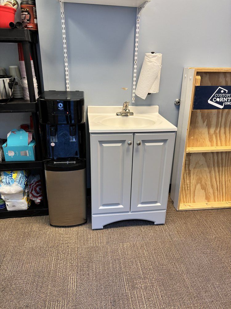 Small break room with a gray sink cabinet, water dispenser, shelving, and a wooden crate shelf.
