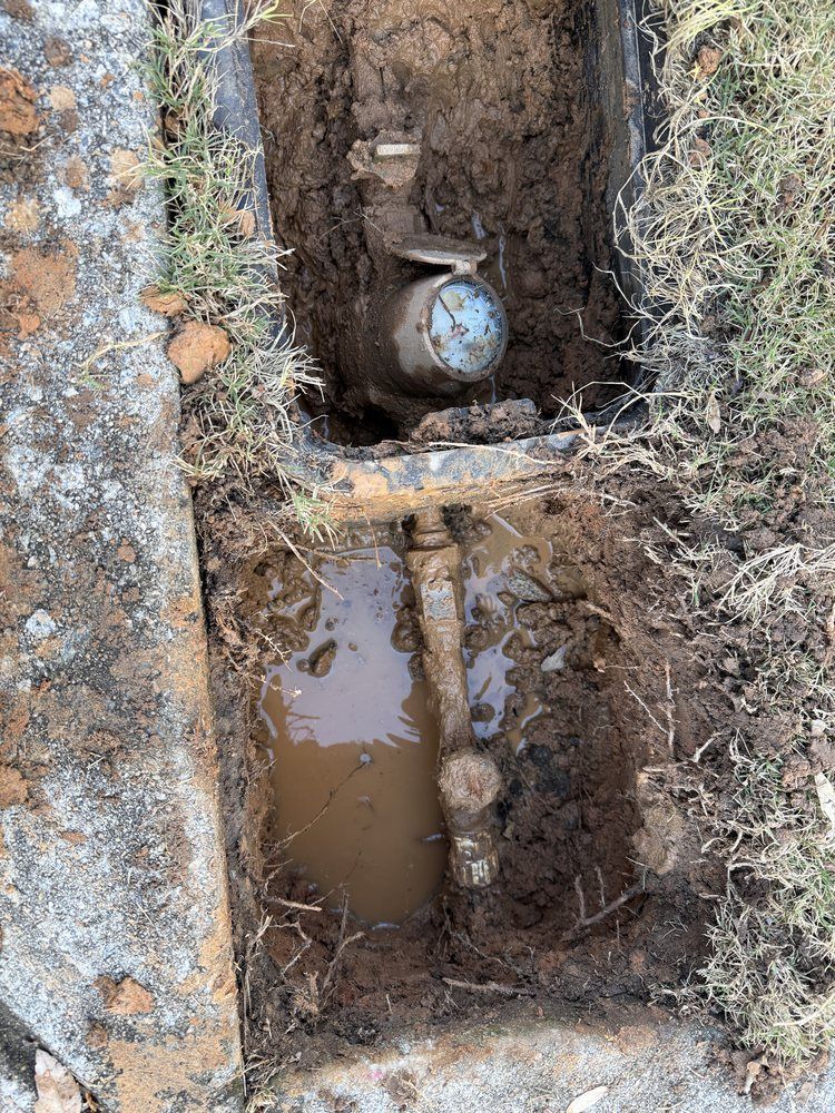 Open utility trench with muddy water, exposed pipe, and a water meter valve.