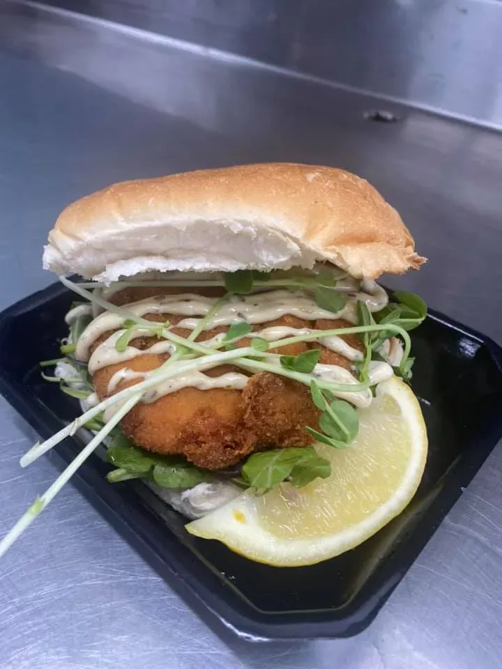 A Fried Chicken Sandwich With a Lemon Wedge on a Black Plate — Stones Oysters & Shed Takeaway in Taree, NSW