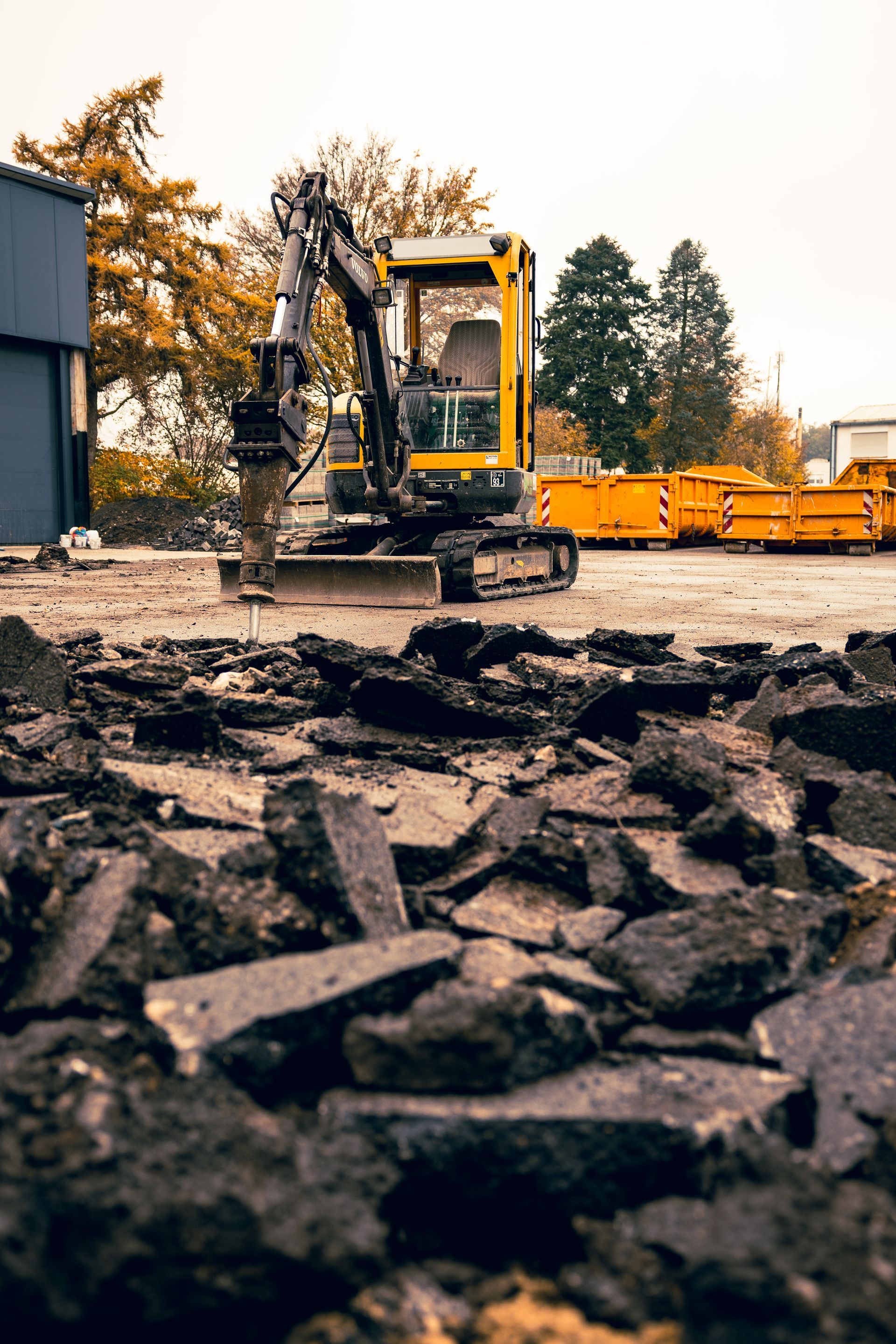 Ein gelber Hydraulikbagger auf einem Schotterplatz, der im Vordergrund Asphalt aufbricht.