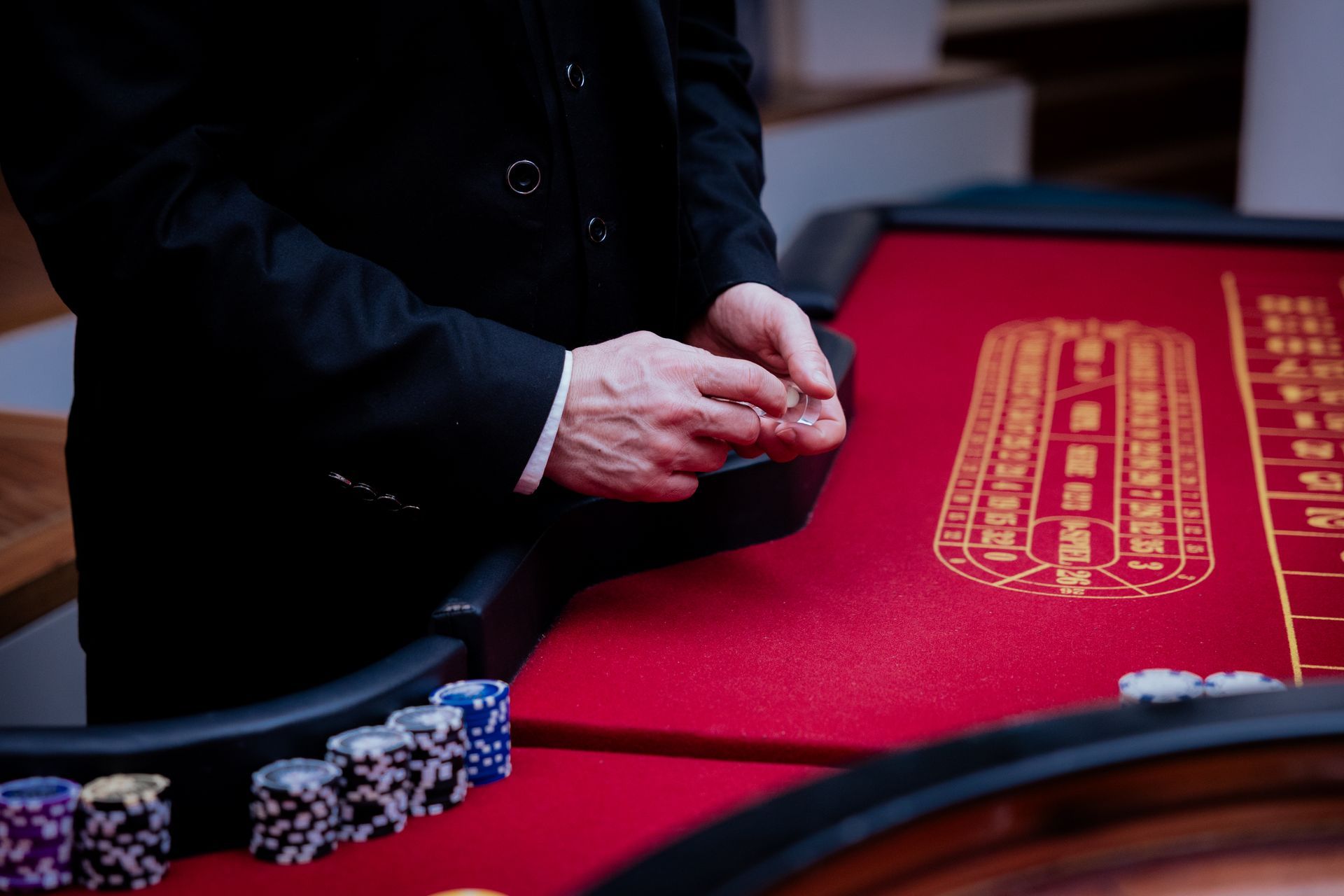 Ein Dealer im Anzug steht an einem roten Casinotisch mit Stapeln von Jetons und einem Roulette-Setzfeld.
