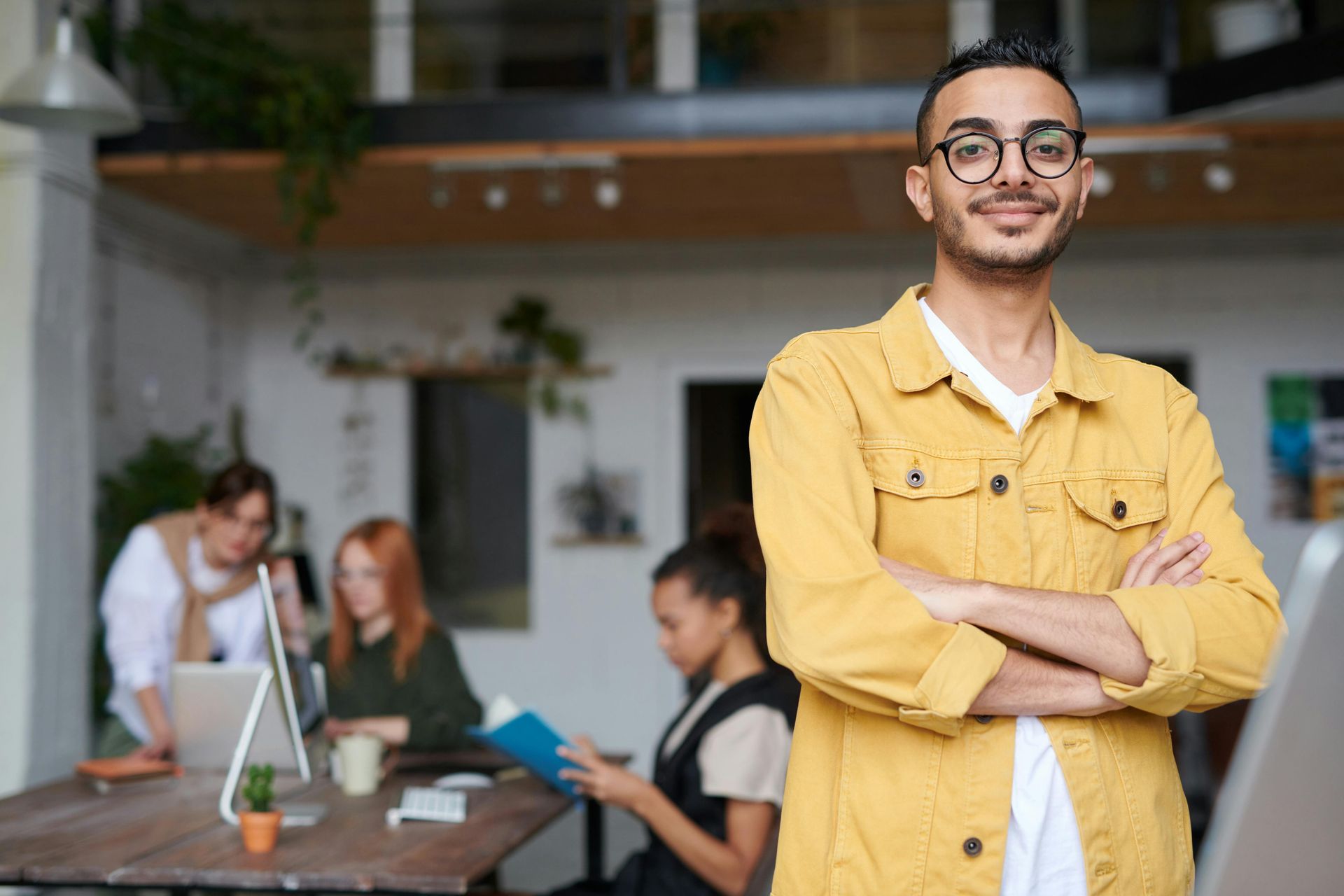 Eine lächelnde Person in einer gelben Jacke steht mit verschränkten Armen in einem Büro, im Hintergrund arbeiten Kollegen.