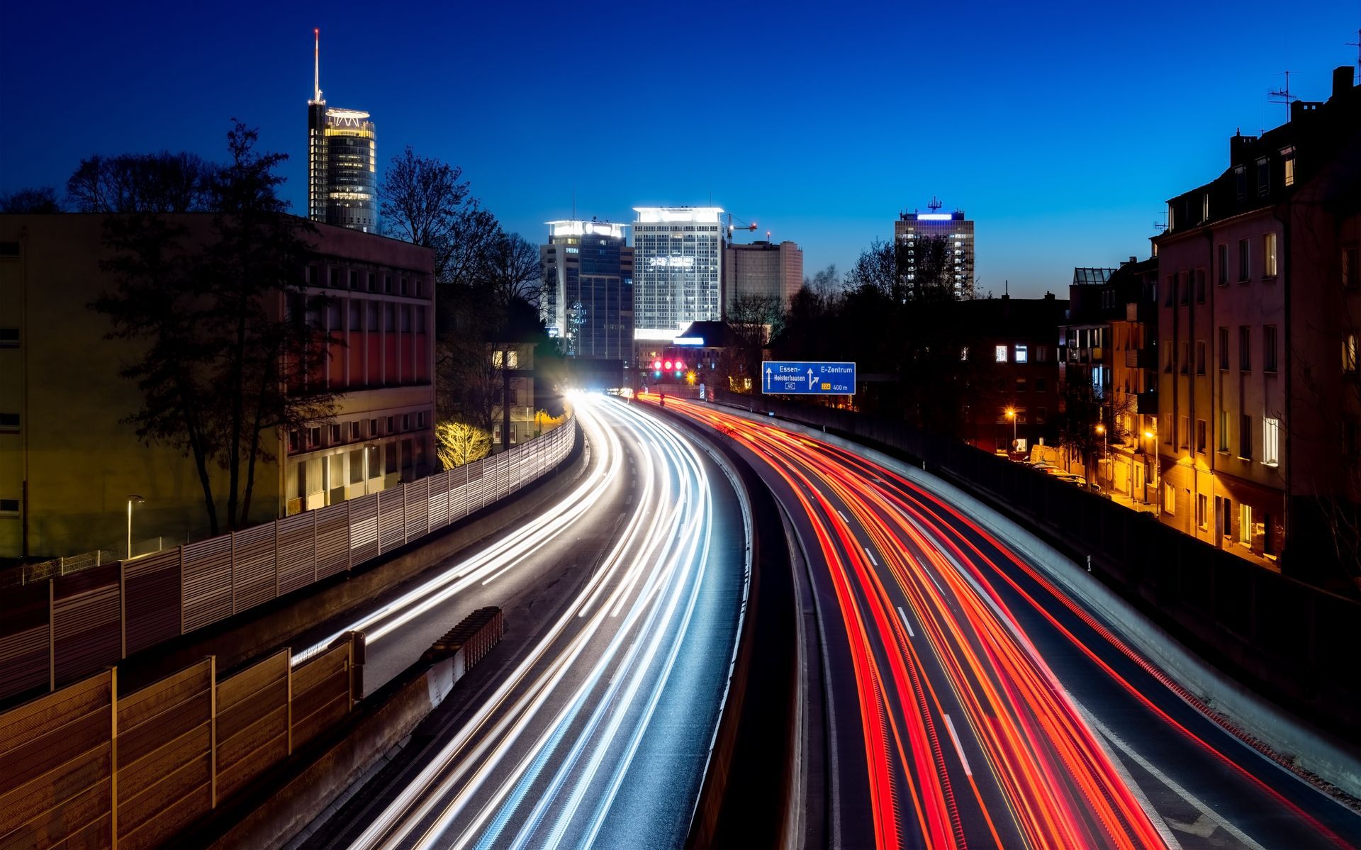 Nachtaufnahme mit Langzeitbelichtung einer befahrenen Autobahn, die weiße und Lichtspuren vor der Skyline einer Stadt zeigt.