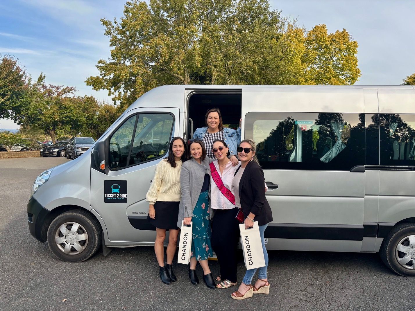 A group of women smiling in front of a Ticket 2 Ride minibus while on a hens day trip.