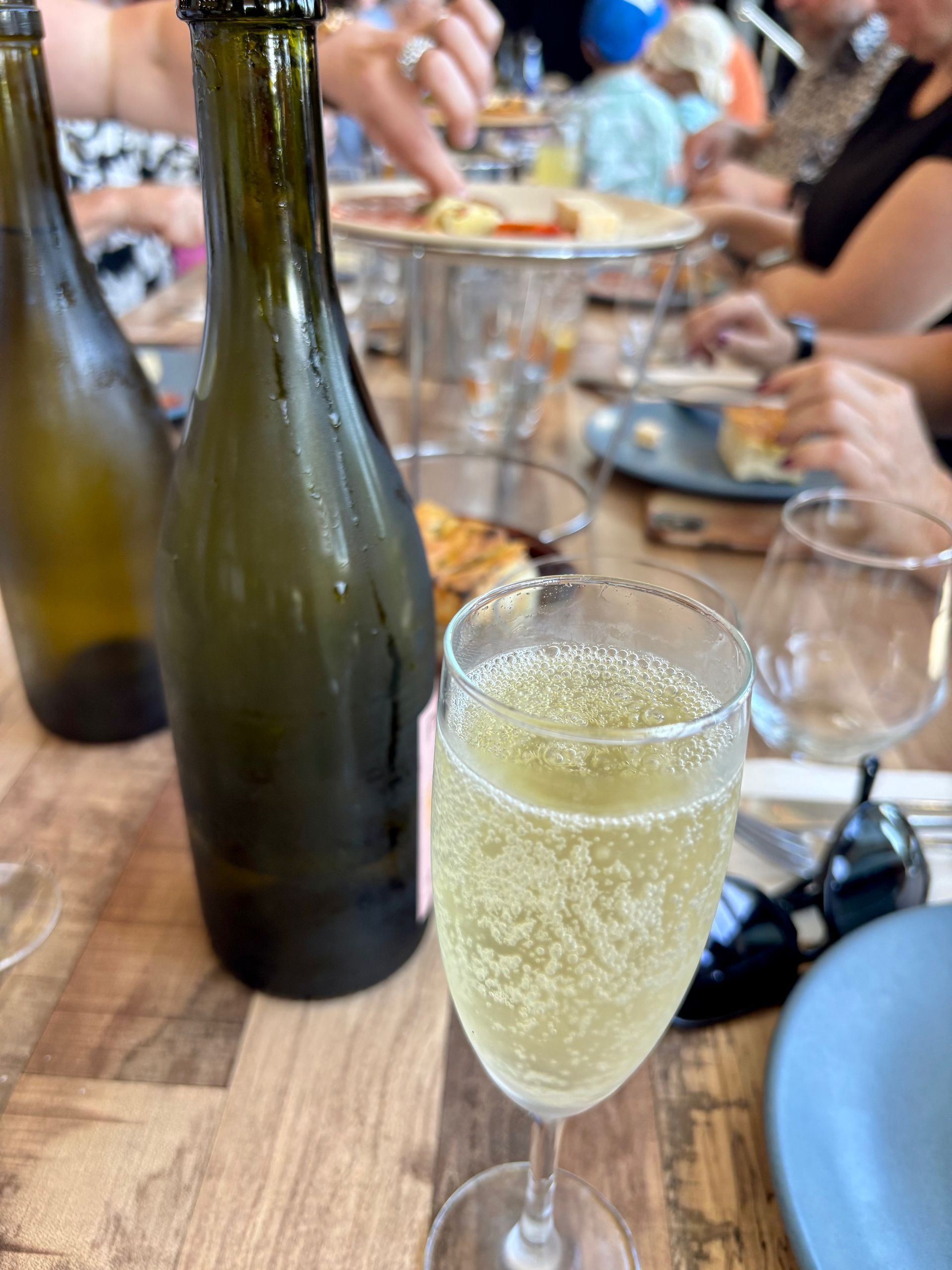 A close up photo of a winery table showing two wine bottles and a glass of bubbles with people enjoying food platters.