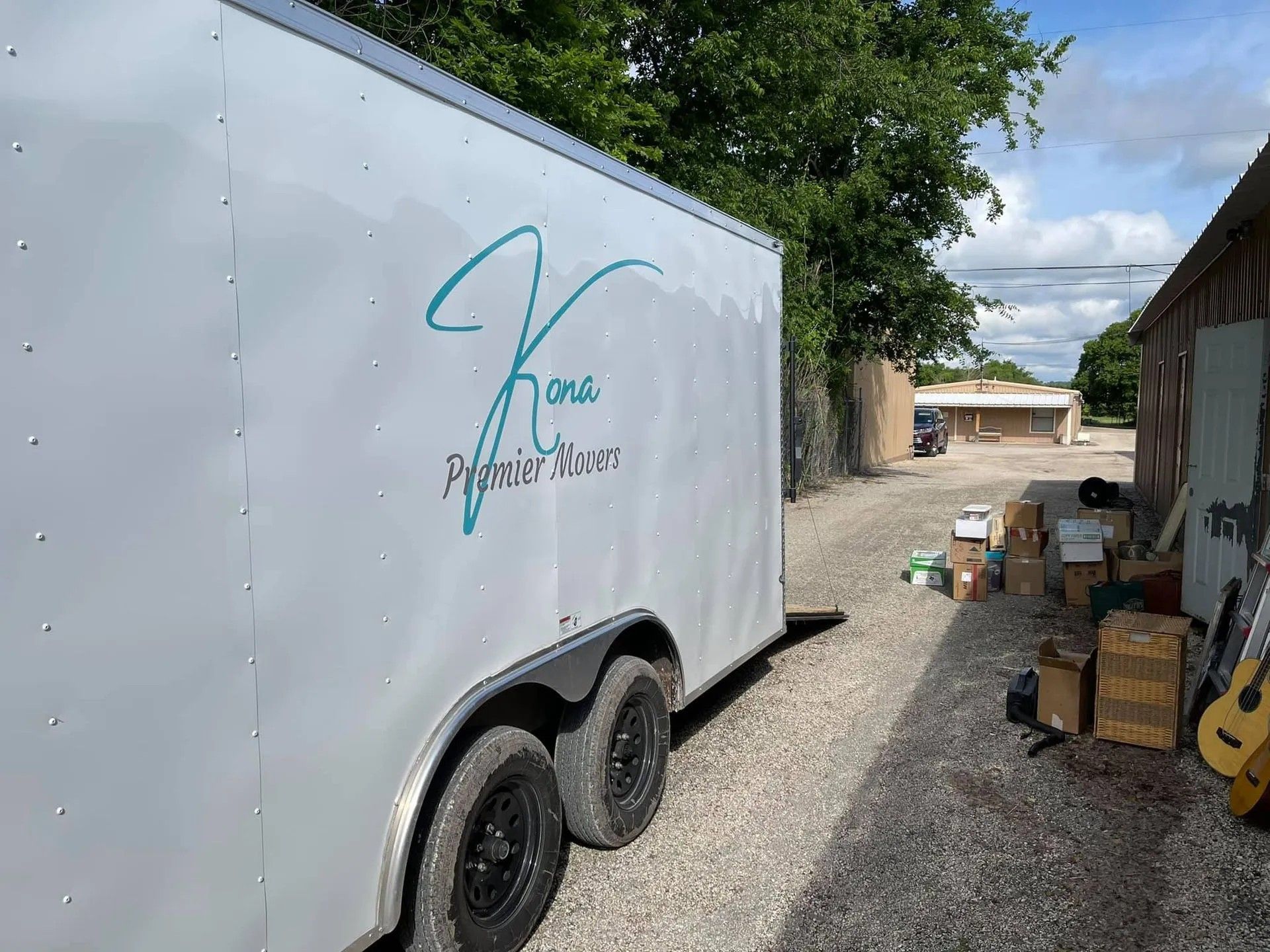 White trailer with teal logo parked on gravel beside a shed and yard clutter under a cloudy sky