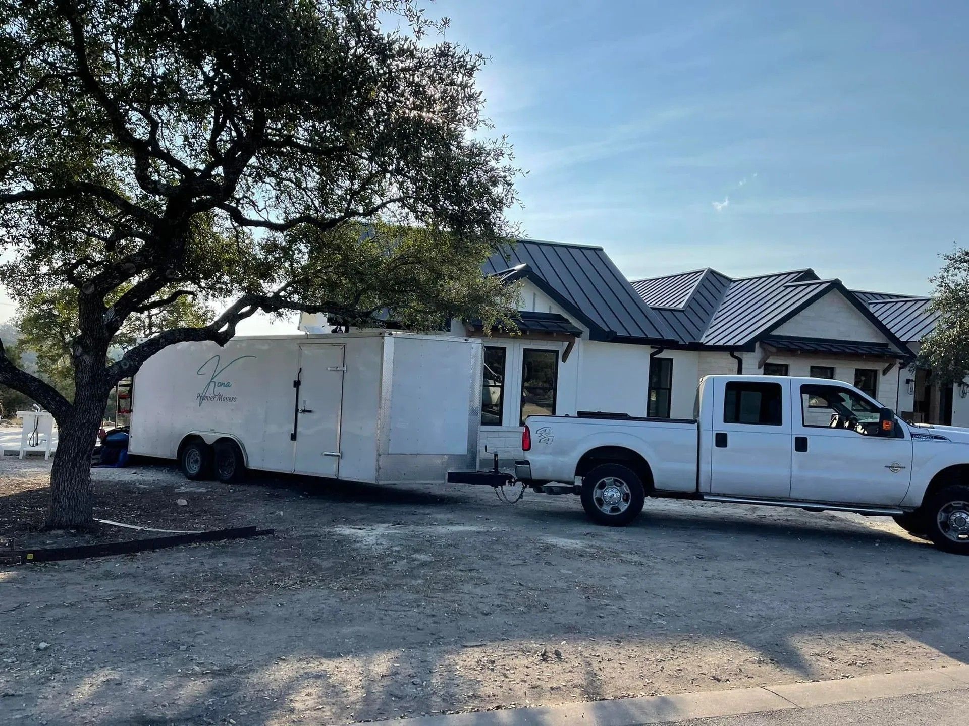 White pickup and trailer parked beside a house under a tree on a dusty lot.