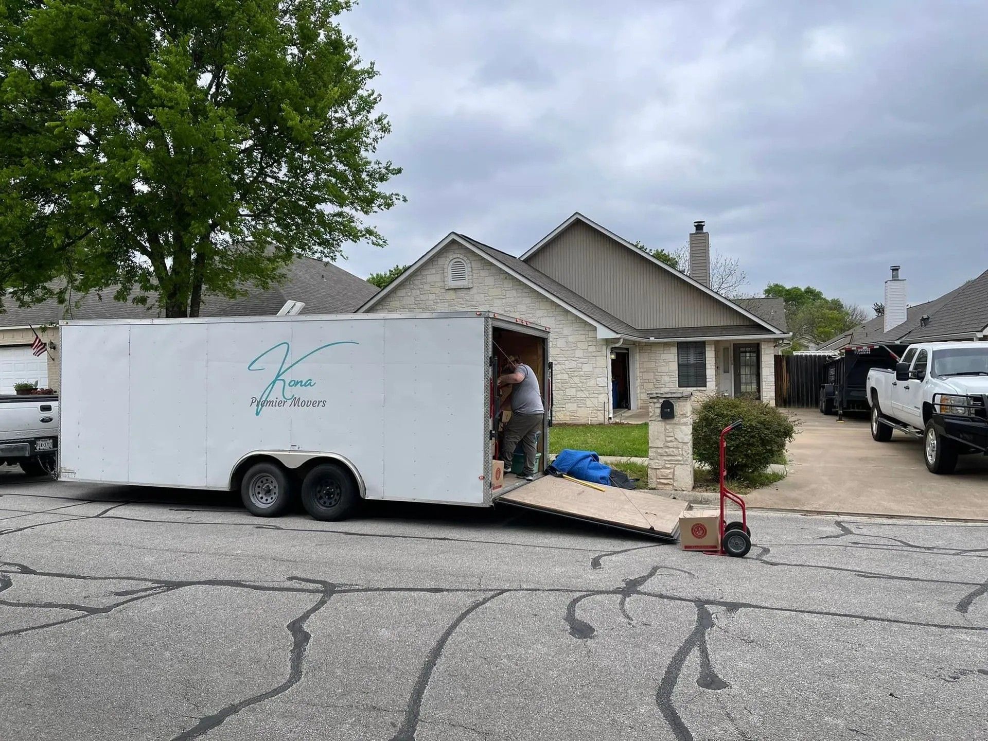 Moving crew unloading a white trailer beside a suburban house with stone exterior and parked vehicles.