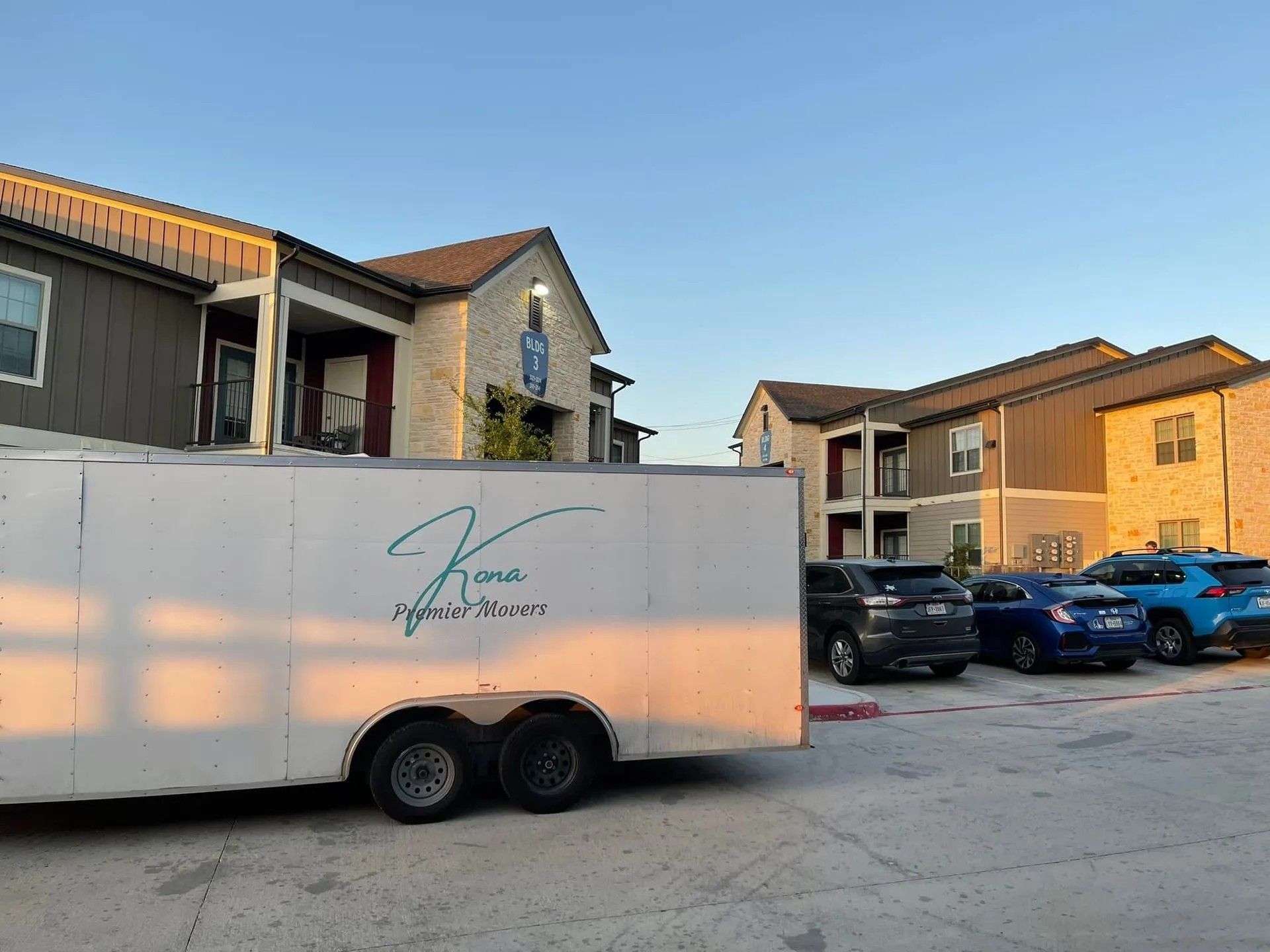 White trailer parked in an apartment lot beside several cars under a clear sky.