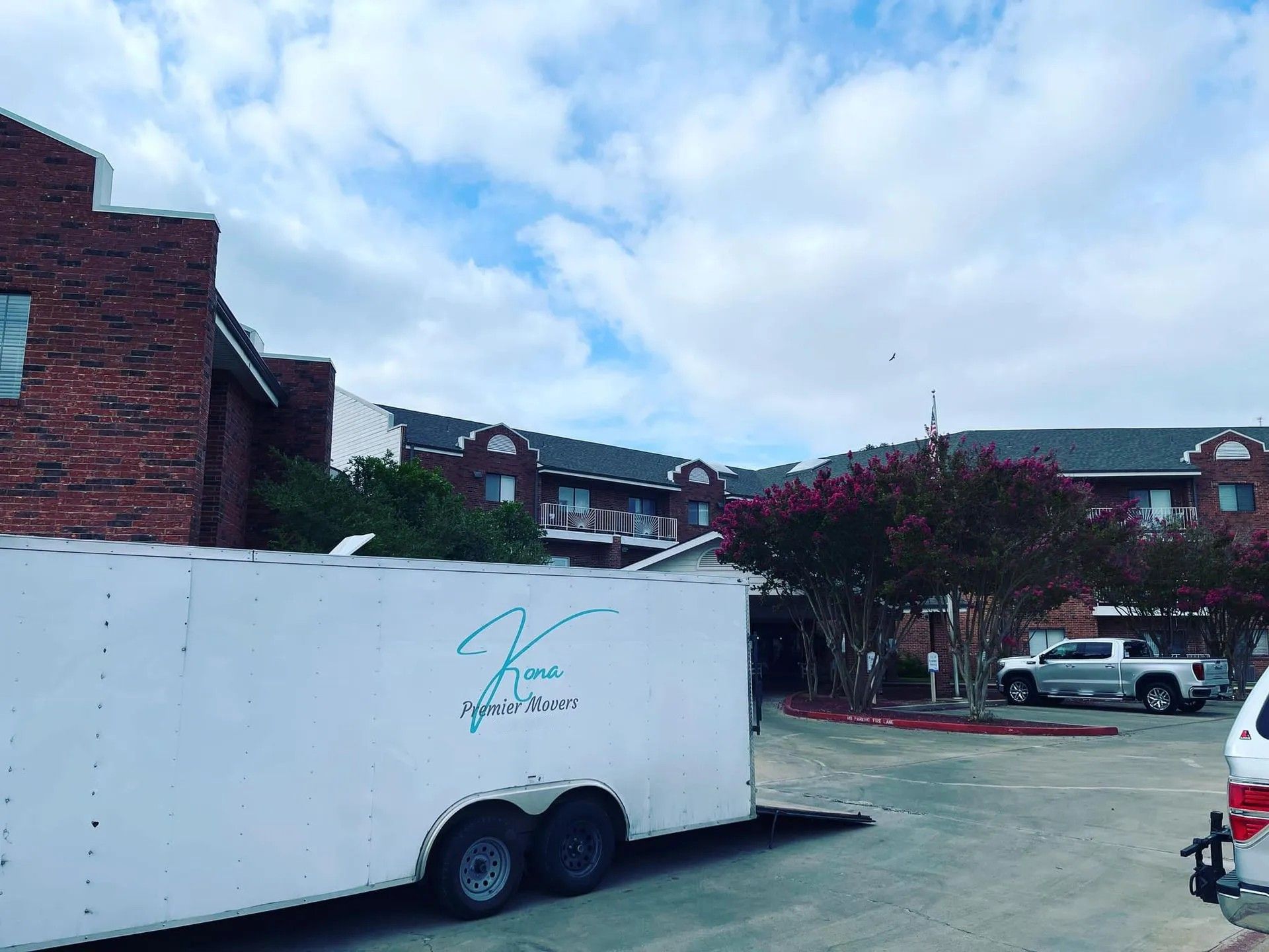 White trailer parked by brick apartments under a cloudy sky, with trees and parked cars nearby